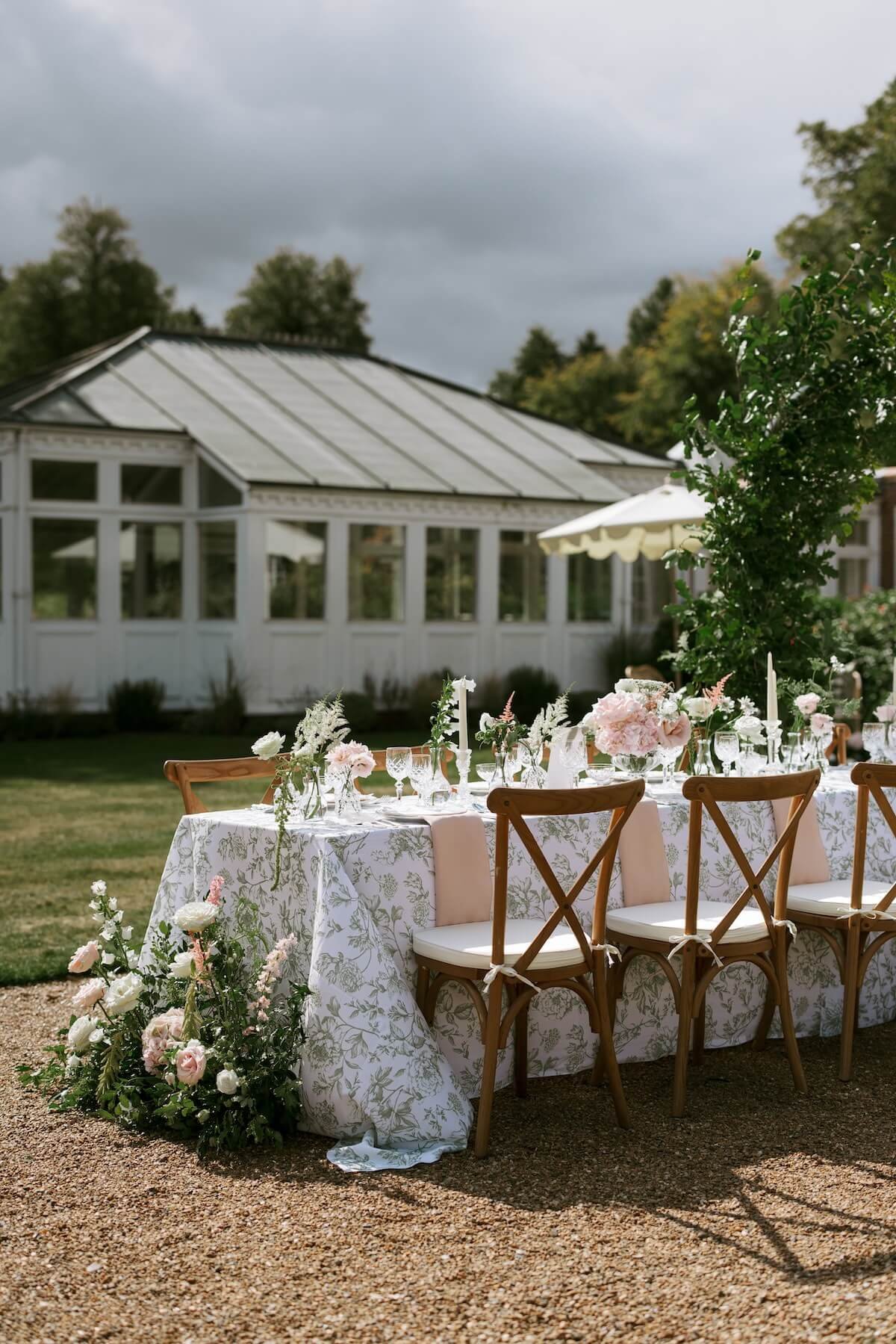 Outdoor wedding or event setup with a long floral table, wooden chairs, and a white building in the background under cloudy sky