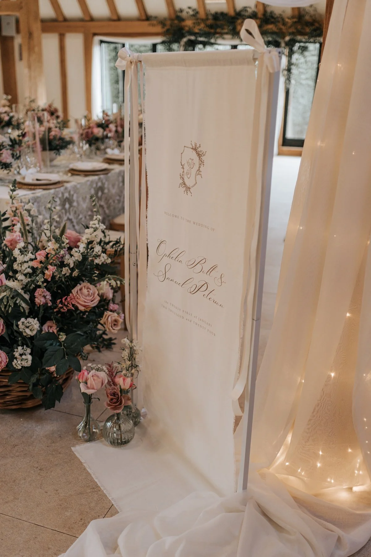 Wedding reception decor with a welcome sign for Ophelia Bell and Samuel Peterson, surrounded by pink and white flowers in vases at a decorated table.