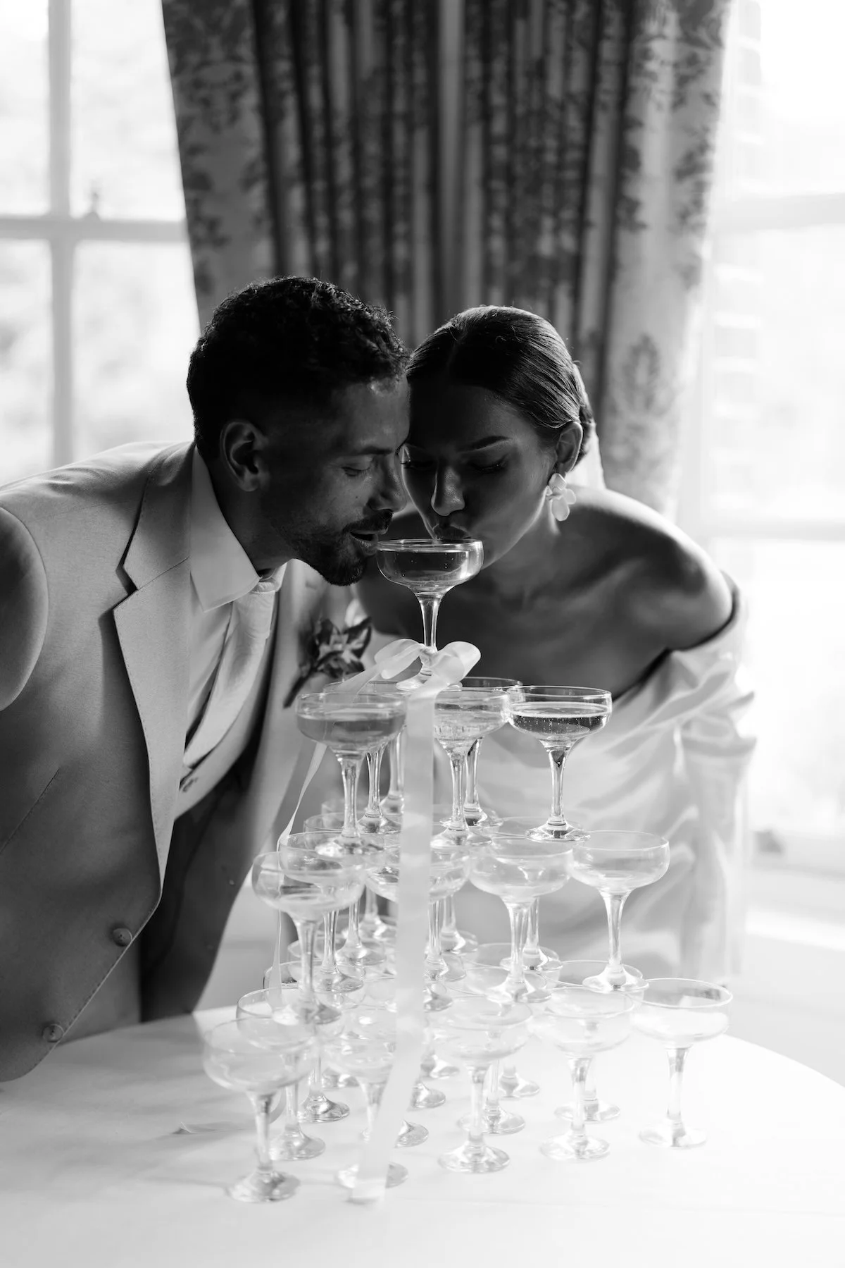 A black and white photo of a man and woman in wedding attire leaning over a pyramid of champagne glasses, drinking from the top glass during a celebration.