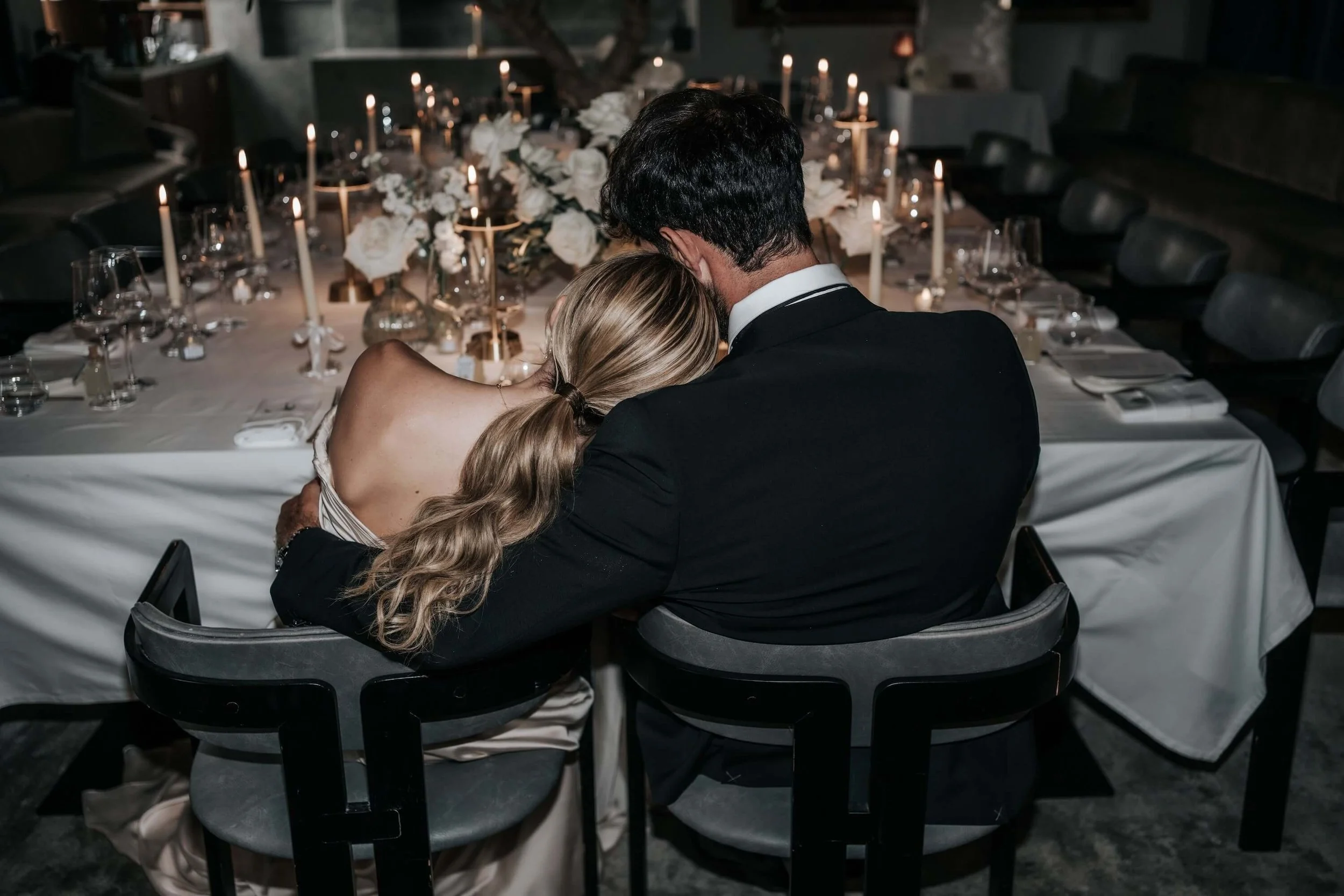 A couple seated at a decorated dining table, embracing each other, in a dimly lit, elegant setting with white flowers and candles.