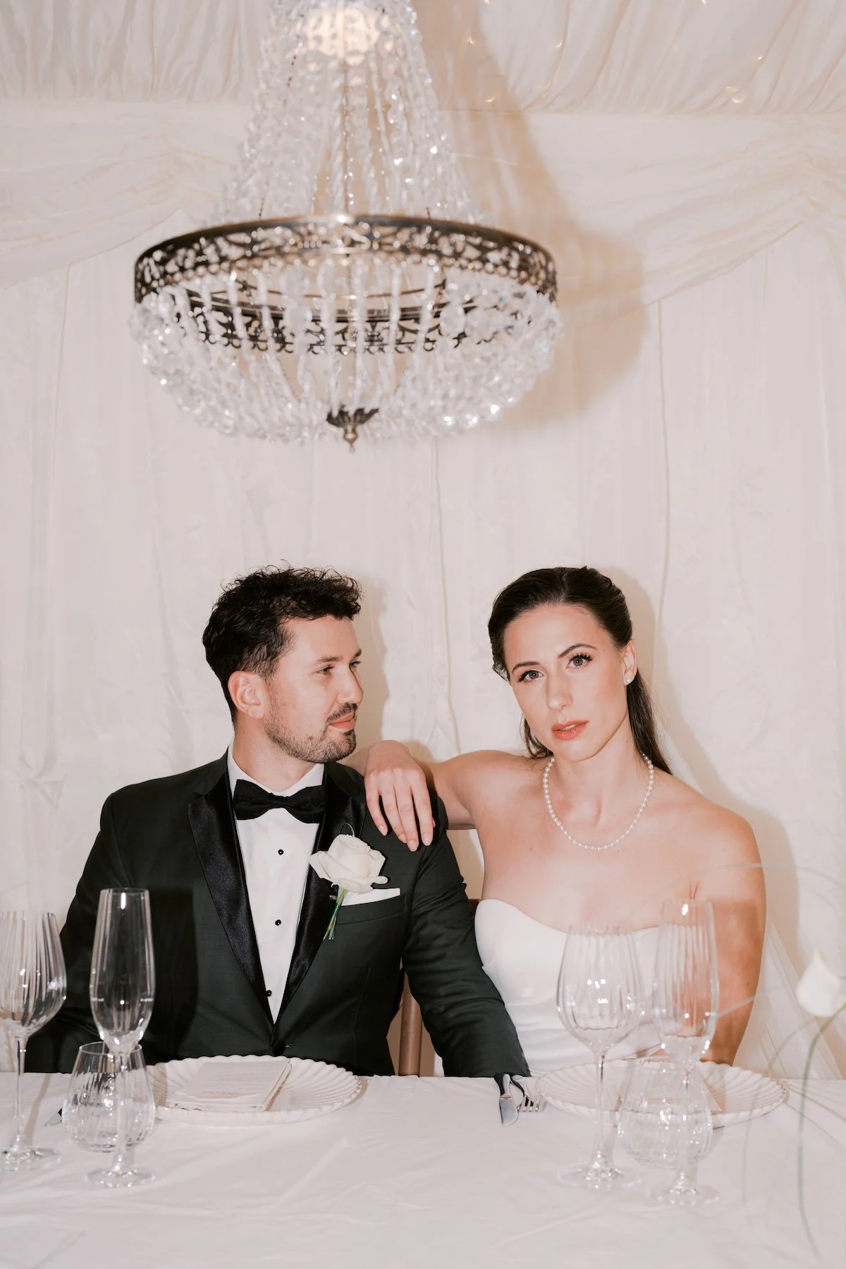 A bride and groom sitting at a wedding reception table, with a chandelier hanging above them. The groom is wearing a black tuxedo with a white boutonniere, and the bride is in a white strapless wedding dress with a pearl necklace. The table has sever