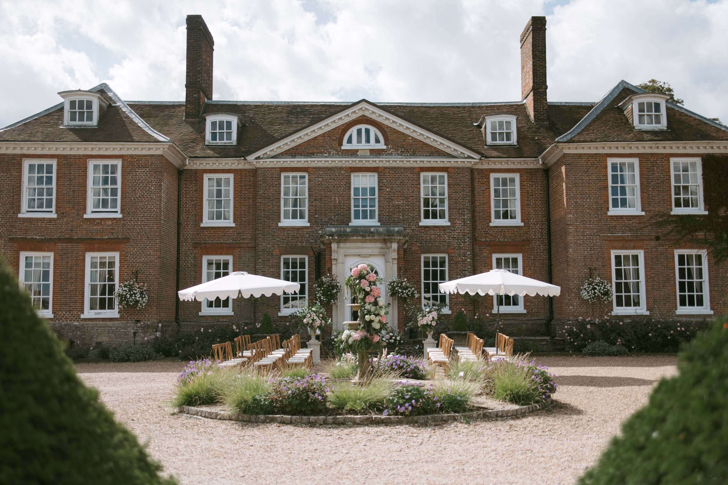 A large brick house with multiple windows and two tall chimneys, decorated with flowers and chairs arranged outdoors, likely for a wedding or outdoor event.