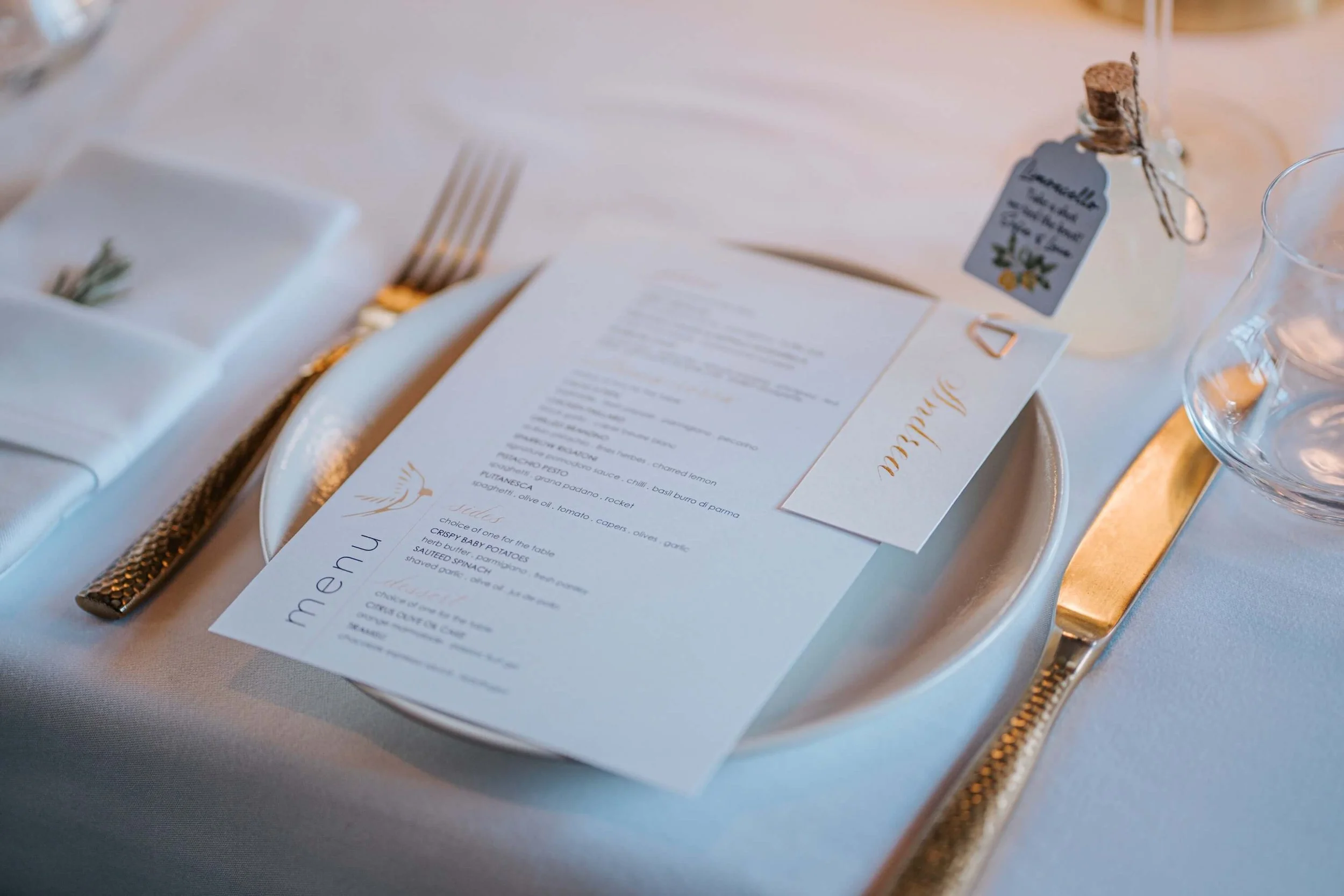 Close-up of a formal dining table setting with a white plate, gold fork, and folded napkin. There is a menu or program on the plate, a small card, and a glass of water. A small decorative tag with a cork and twine is visible, indicating an elegant ev