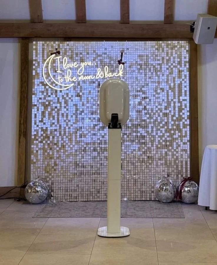 A ceramic hand sanitizer dispenser stand in front of a wall decorated with small, shimmering tiles. On the wall, there is a neon sign with the text 'I love you to the moon & back' and a drawing of a crescent moon. Two mirrored spheres and a white air