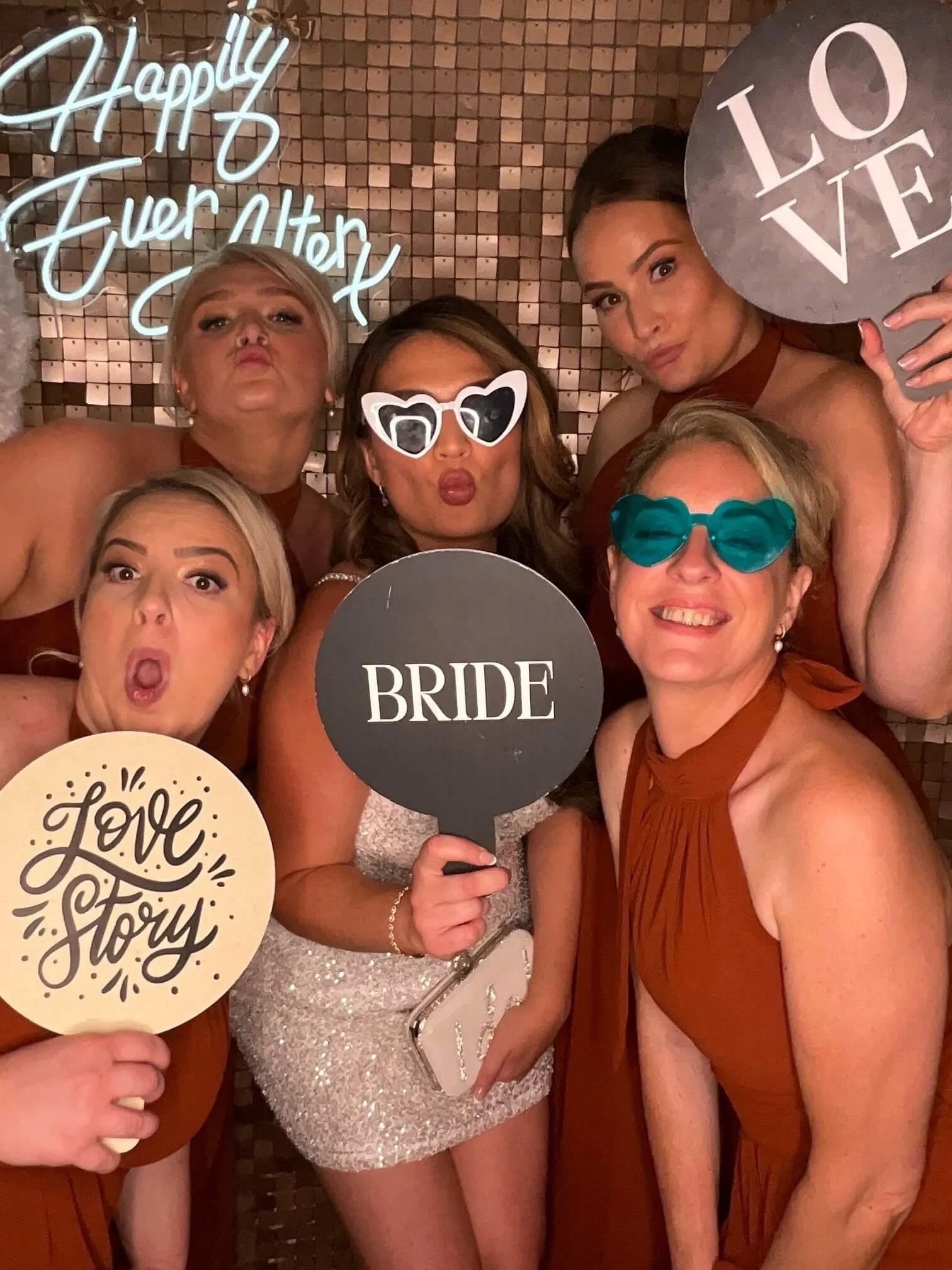 Group of five women at a celebration, holding signs that say 'Love Story' and 'Bride,' wearing party accessories like sunglasses and holding drinks, posing in front of a decorative background.