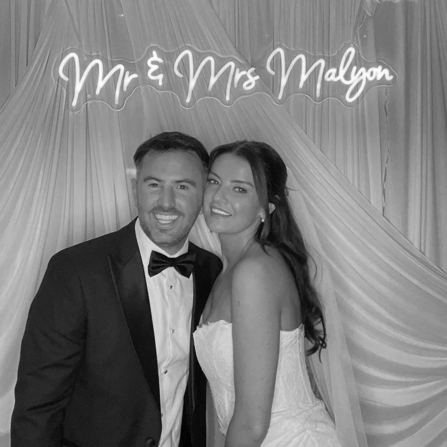 A newlywed couple, a man in a tuxedo and a woman in a wedding dress, smiling and posing for a photo in front of a draped backdrop with a neon sign that says 'Mr & Mrs Malyon'.