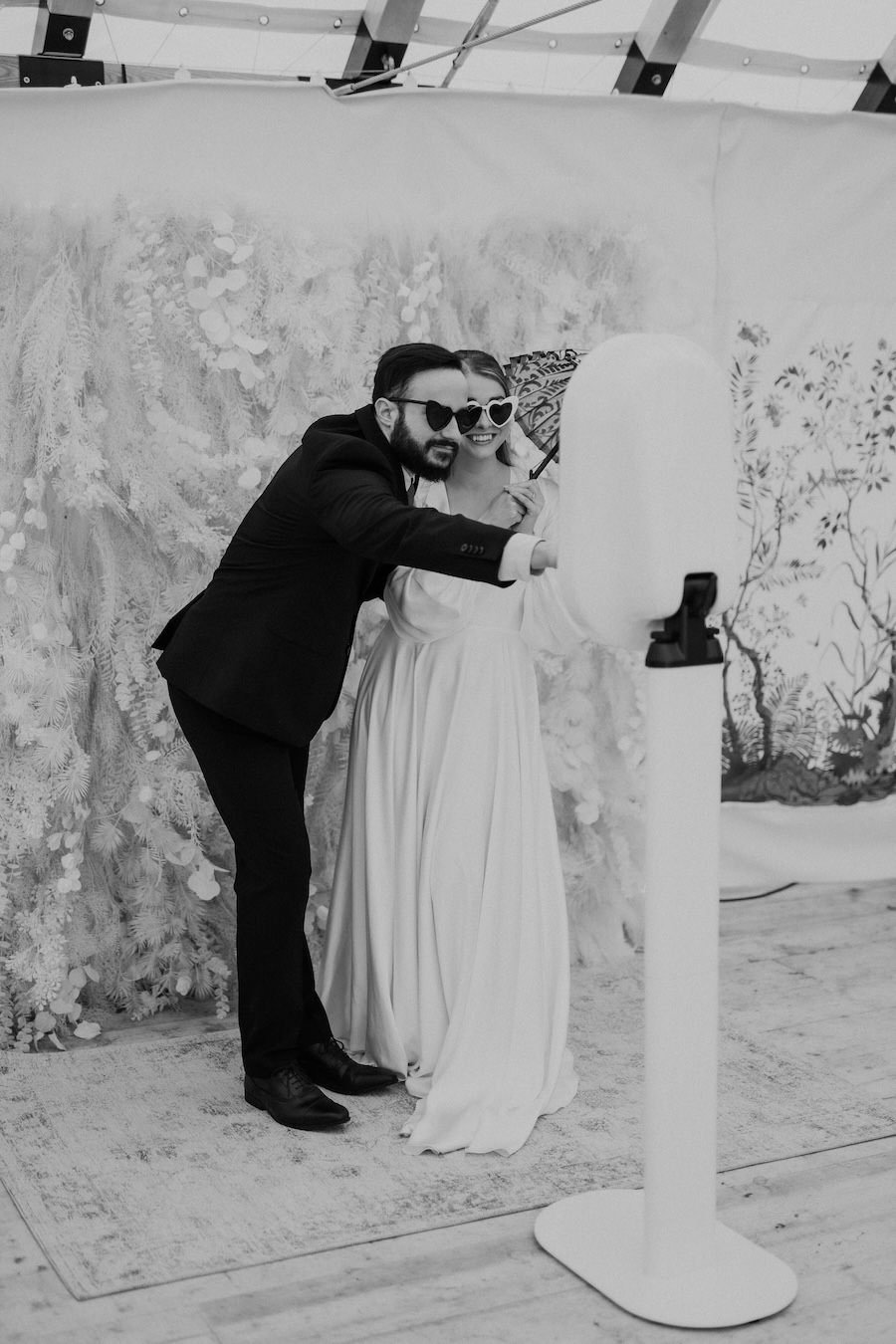 A couple dressed in wedding attire taking a selfie with a selfie stick at a wedding celebration, both wearing sunglasses, with a floral backdrop.
