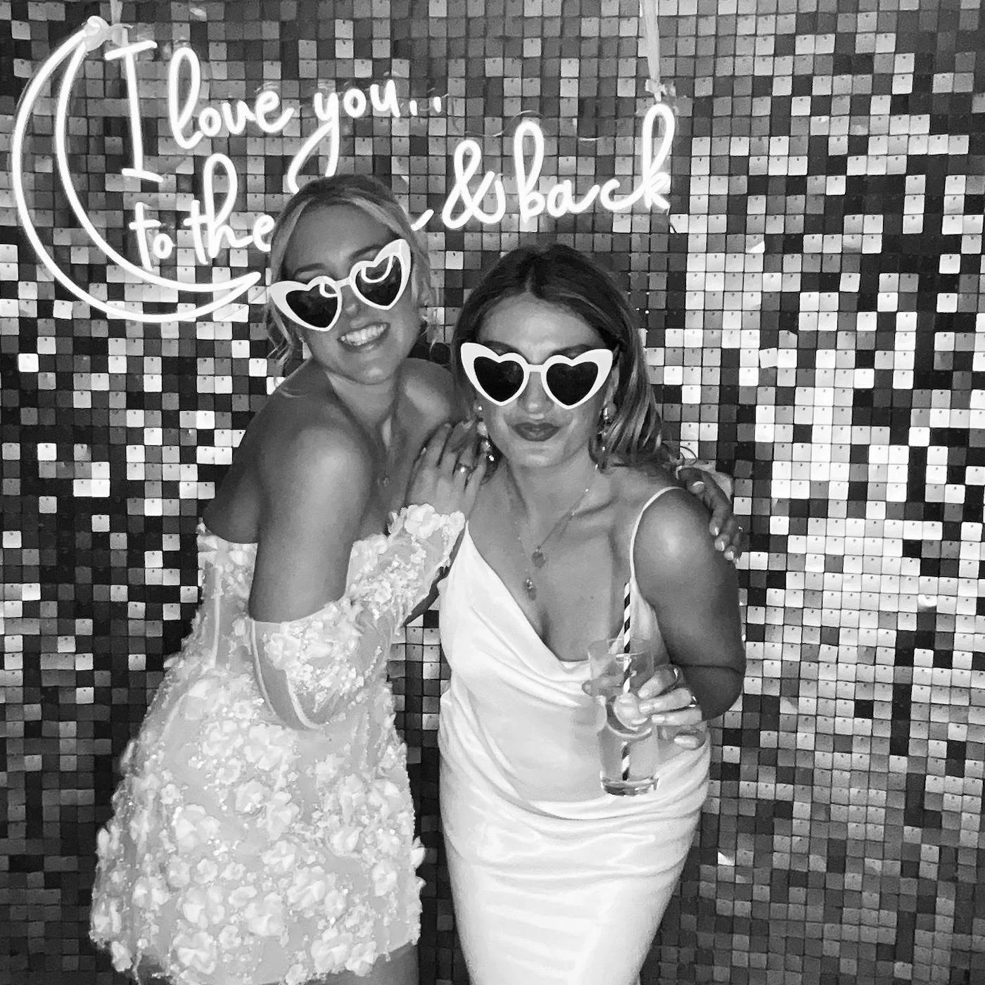 Two women in dresses and sunglasses posing together at a party in front of a mosaic wall with a neon sign that says "I love you to the moon and back".