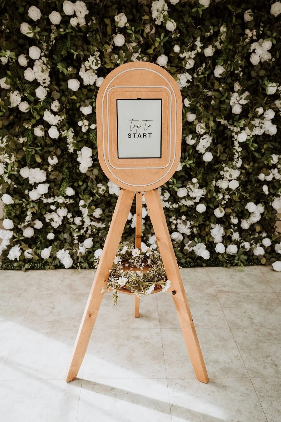 A wooden photo booth with a tablet displaying the words 'Tap to start,' standing in front of a backdrop of white flowers and greenery, with a small flower arrangement hanging from the easel.