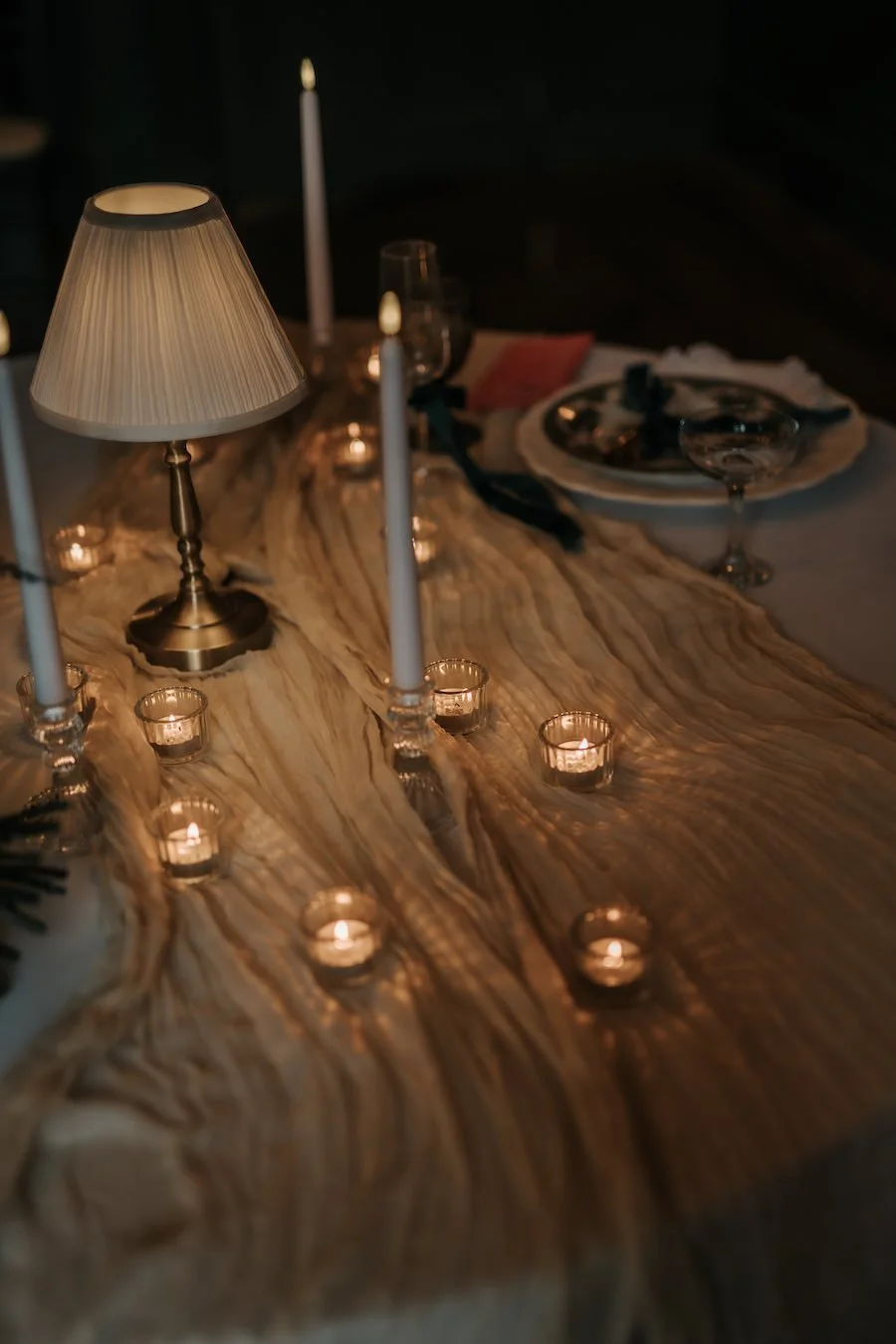 A dimly lit dinner table with candles and a small table lamp, set with plates, glasses, and napkins.