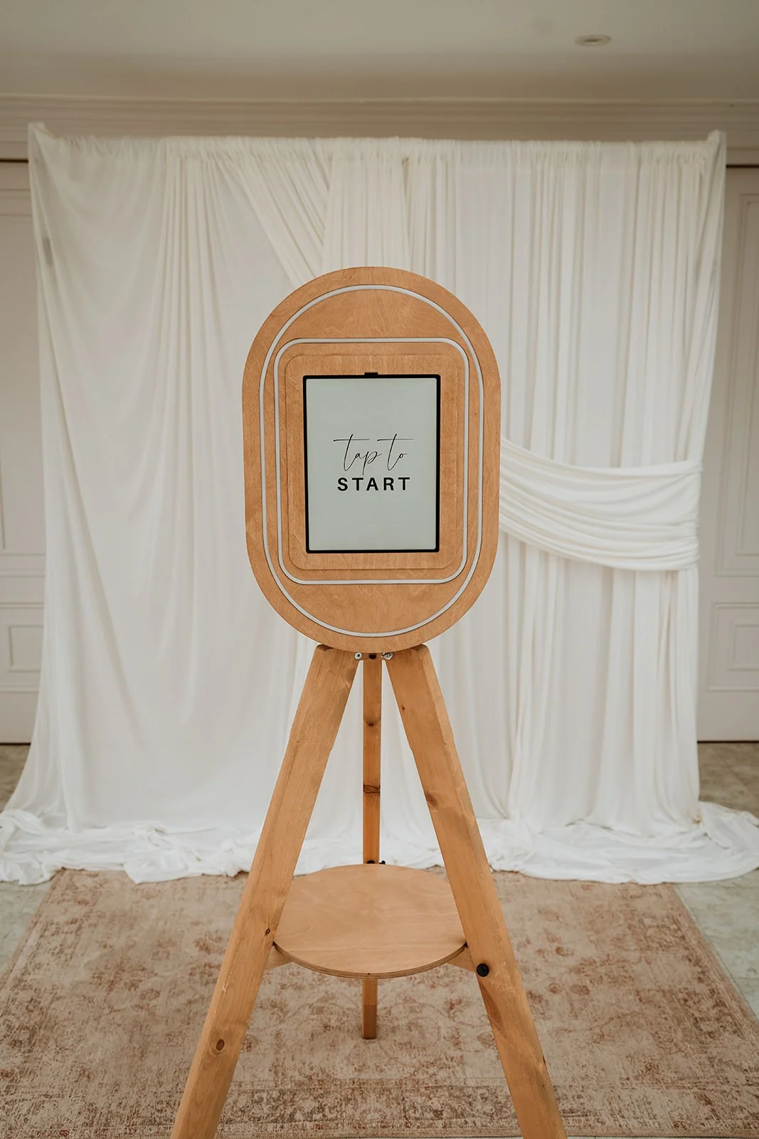 A wooden photo booth with a screen that says 'tap to start' stands on a tripod in front of white draped fabric.