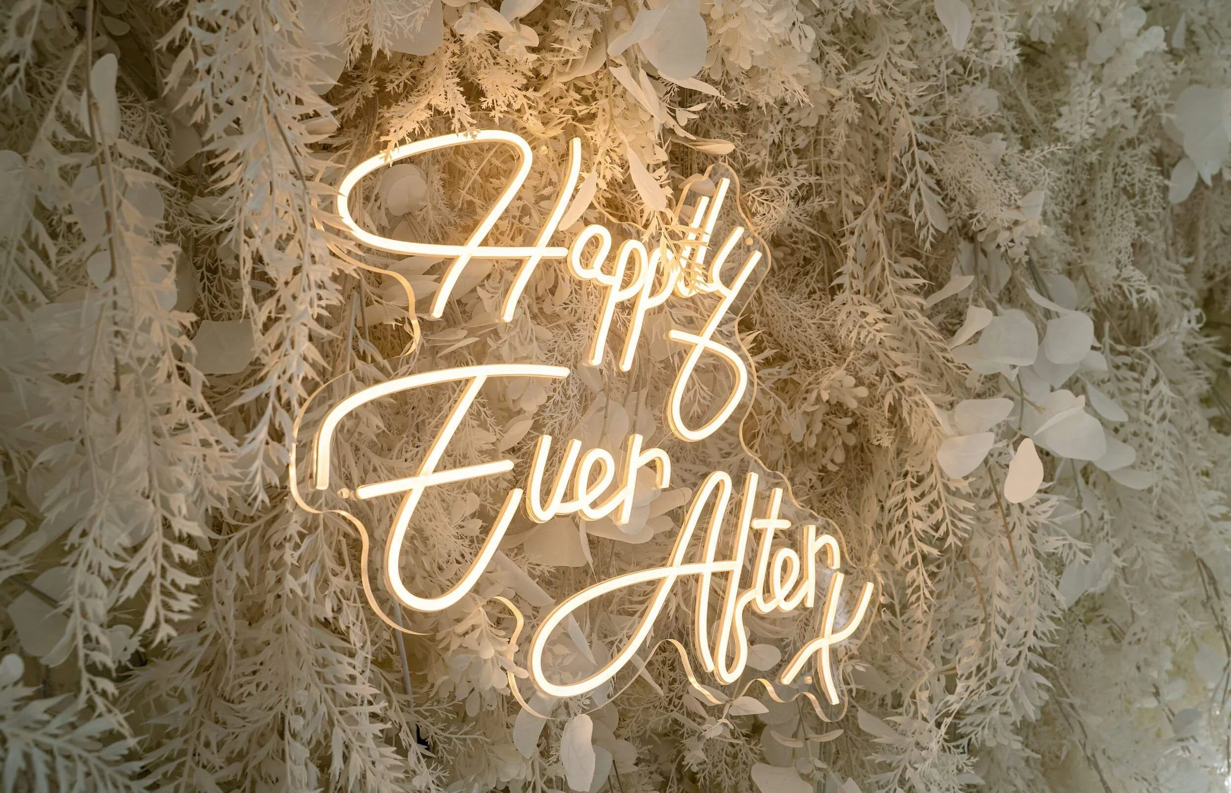 A decorative wall with white faux flowers and leaves, featuring a neon sign that says 'Happy You' in cursive letters.