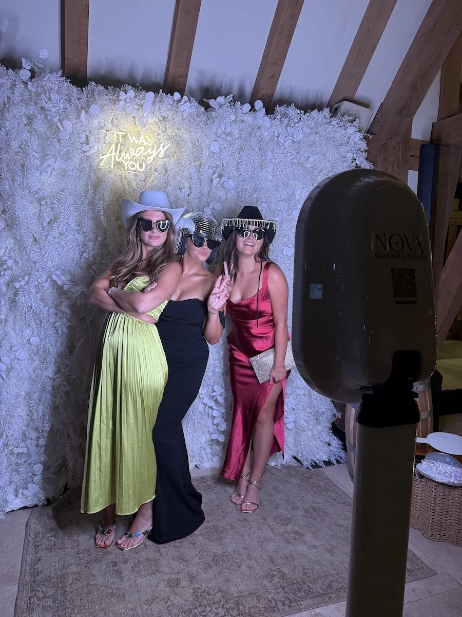 Three women in stylish dresses and large hats taking a photo in front of a floral backdrop with a neon sign that says 'It was always you'. They are wearing sunglasses, and one woman is making a peace sign. There is a large camera or photo booth in th
