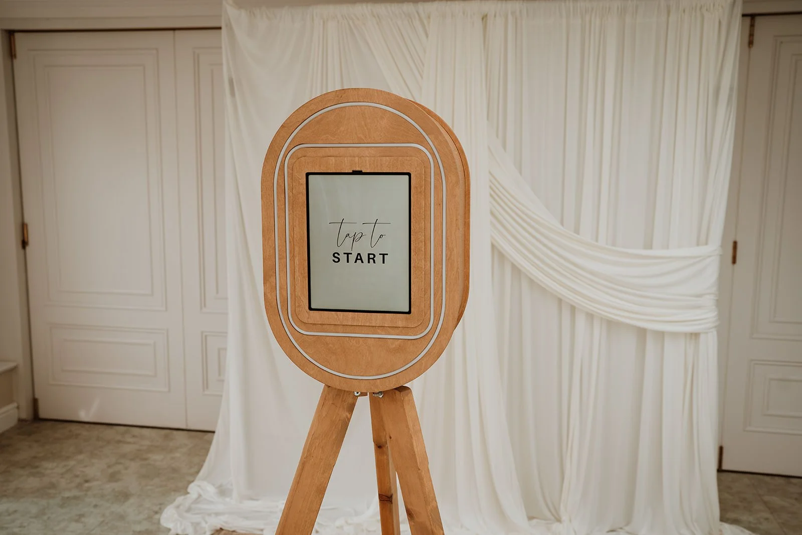 photo booth with a digital screen that says 'Tap to START' in an elegant font. The background features white draped curtains and closed white doors.
