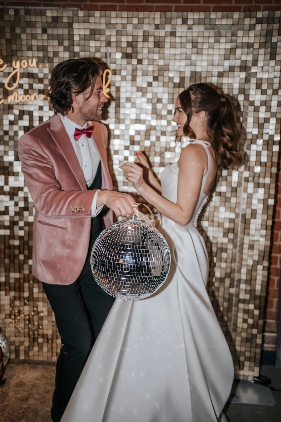A man and woman dancing at a celebration, with the man holding a disco ball, against a sparkly tiled wall backdrop.