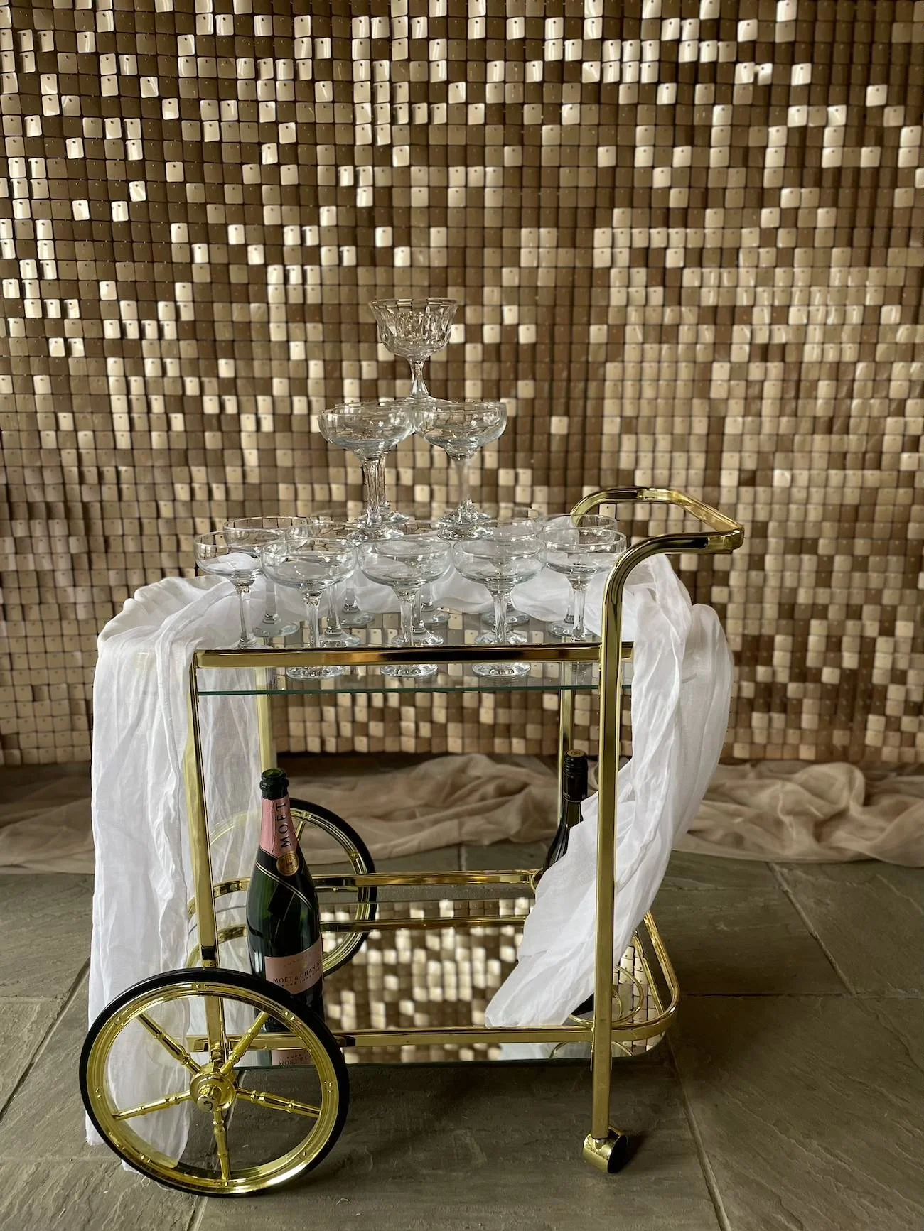 A gold bar cart with glassware and a bottle of champagne. The cart is draped with white fabric, and there is a decorative backdrop made of small golden tiles.
