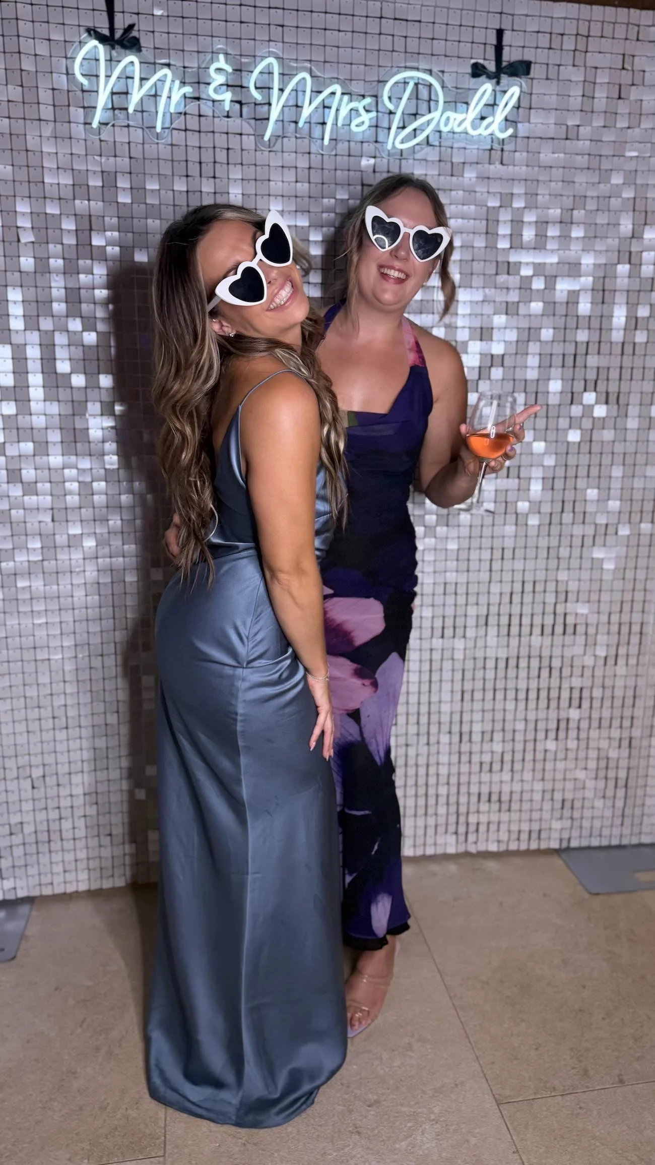 Two women wearing sunglasses with heart-shaped lenses, smiling, and standing in front of a sign that reads 'Mr. & Mrs. Dodd'. One woman is holding a glass of rosé wine and wearing a silky, strapless gown, while the other is wearing a colorful dress.