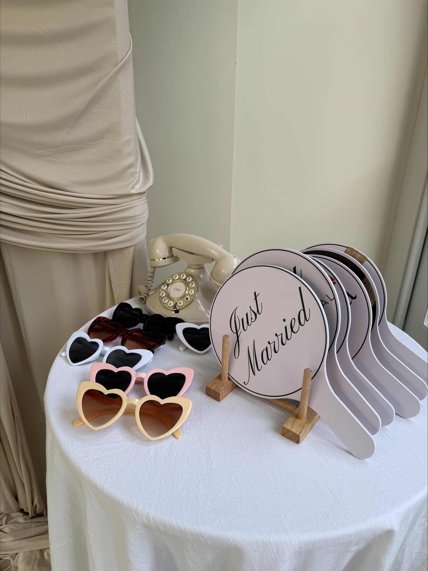Table with accessories for a wedding or bridal event, including sunglasses in various styles and colors, round signs that say "Just Married," and a vintage white rotary telephone.