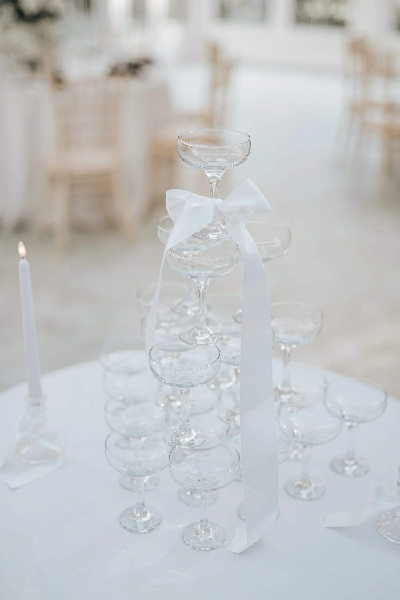 Empty champagne glasses stacked in a pyramid with a white ribbon tied around the top glass, on a white tablecloth in a bright, elegant setting.