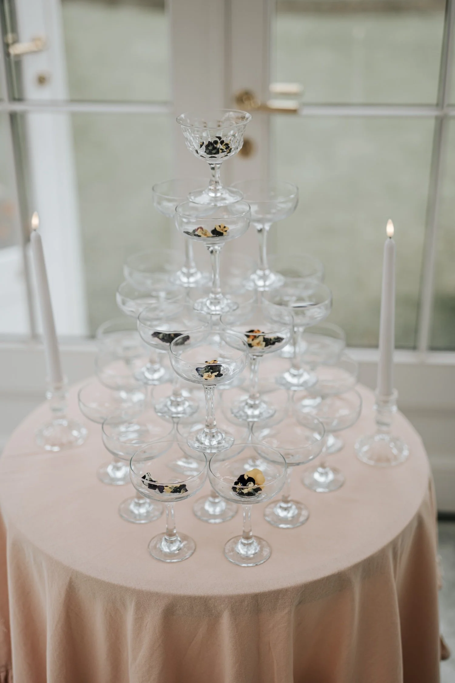 A tower of empty champagne coupe glasses with black and gold confetti inside, arranged on a round table with a pink tablecloth, near lit white candles, in front of a window.