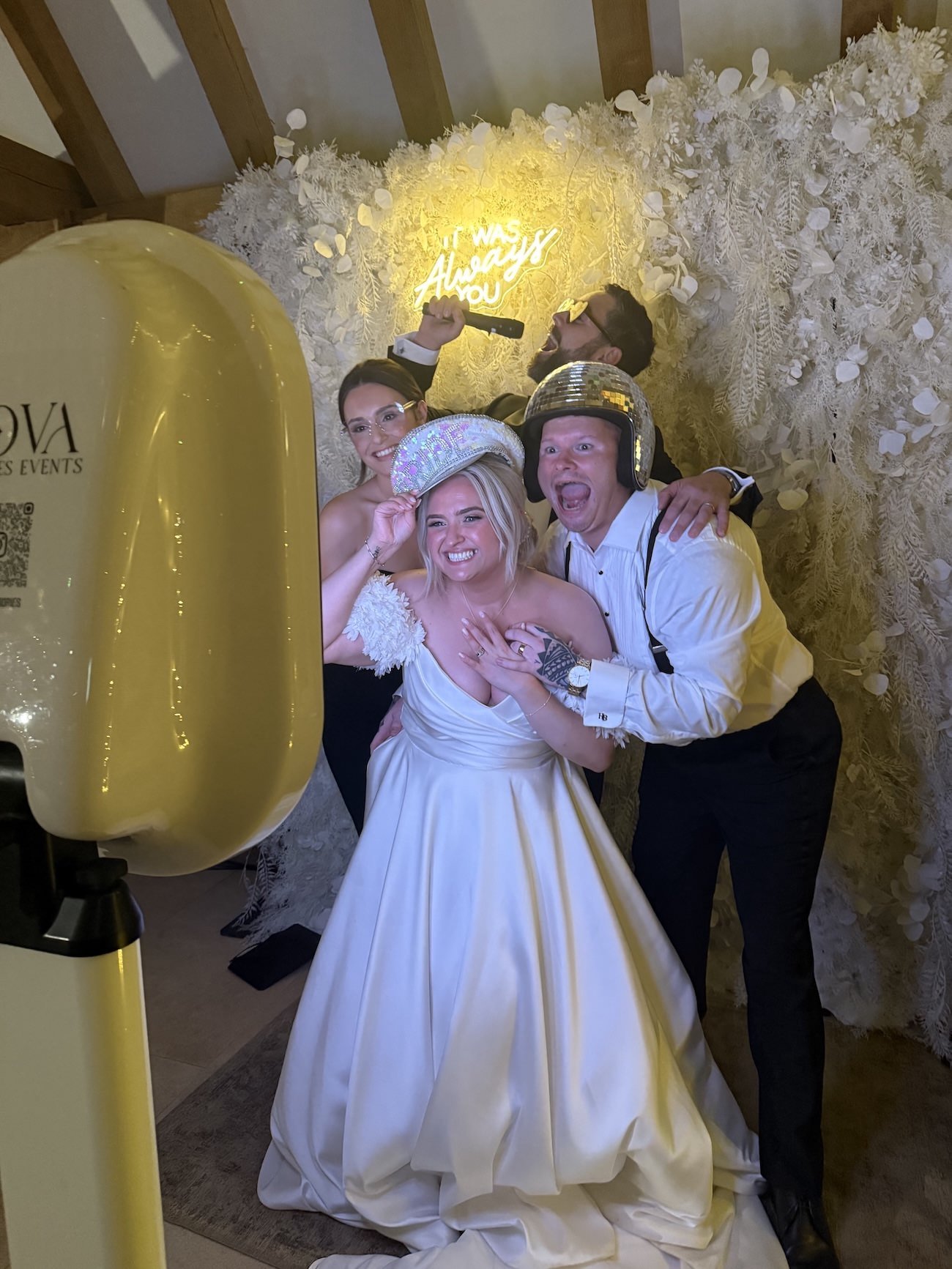 Group of four people celebrating at a wedding photo booth, with a white floral backdrop and a neon sign that says "It Was Always You." One woman in a wedding dress holds a sparkly birthday hat, while a man wearing a helmet and a woman in glasses hold props and make expressive faces.
