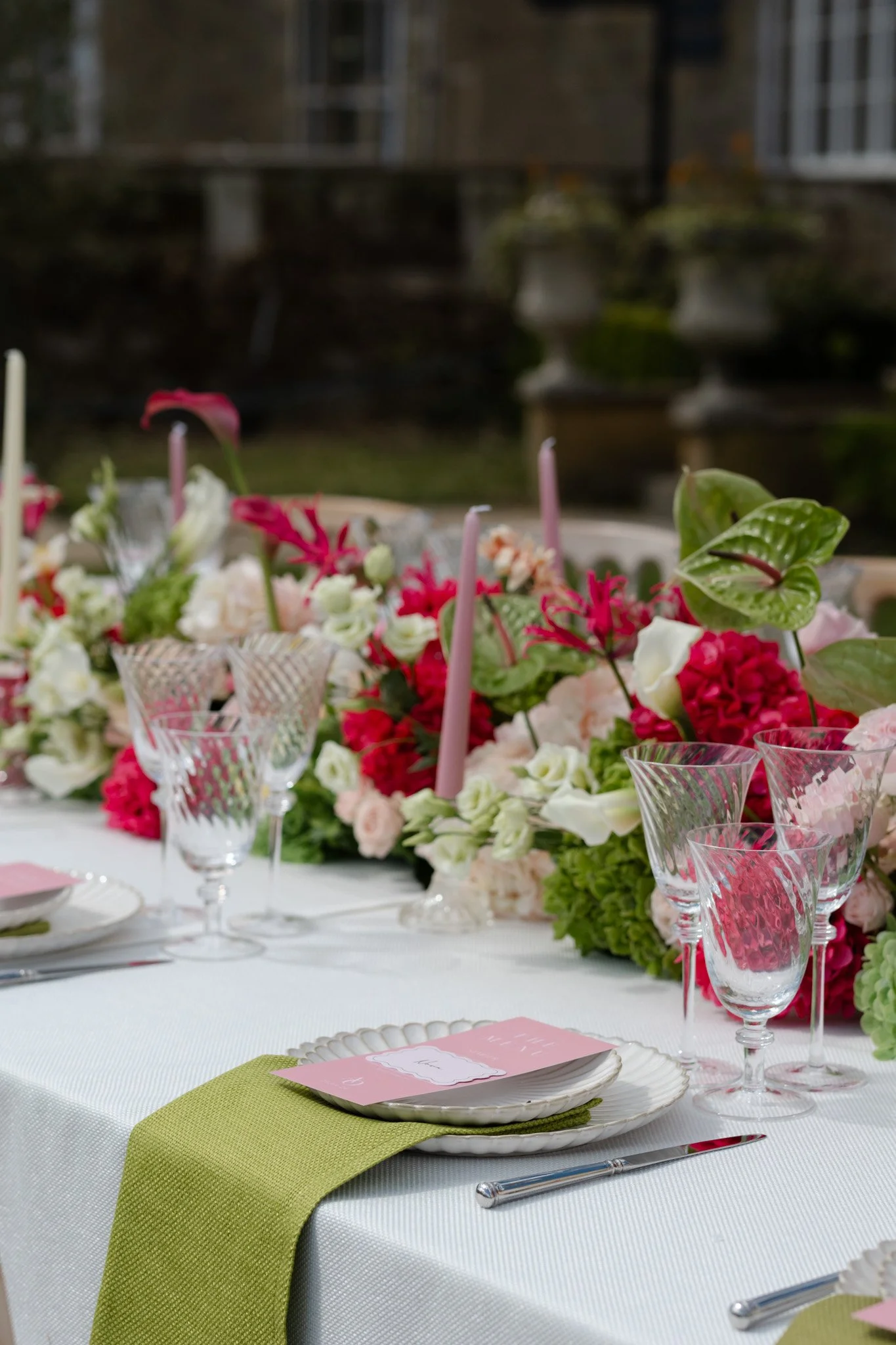 A decorated outdoor table set for an event, featuring a floral centerpiece with pink, white, and red flowers, pink candles, clear glassware with patterned grips, white plates, silverware, and pink place cards, with a building and garden in the backgr