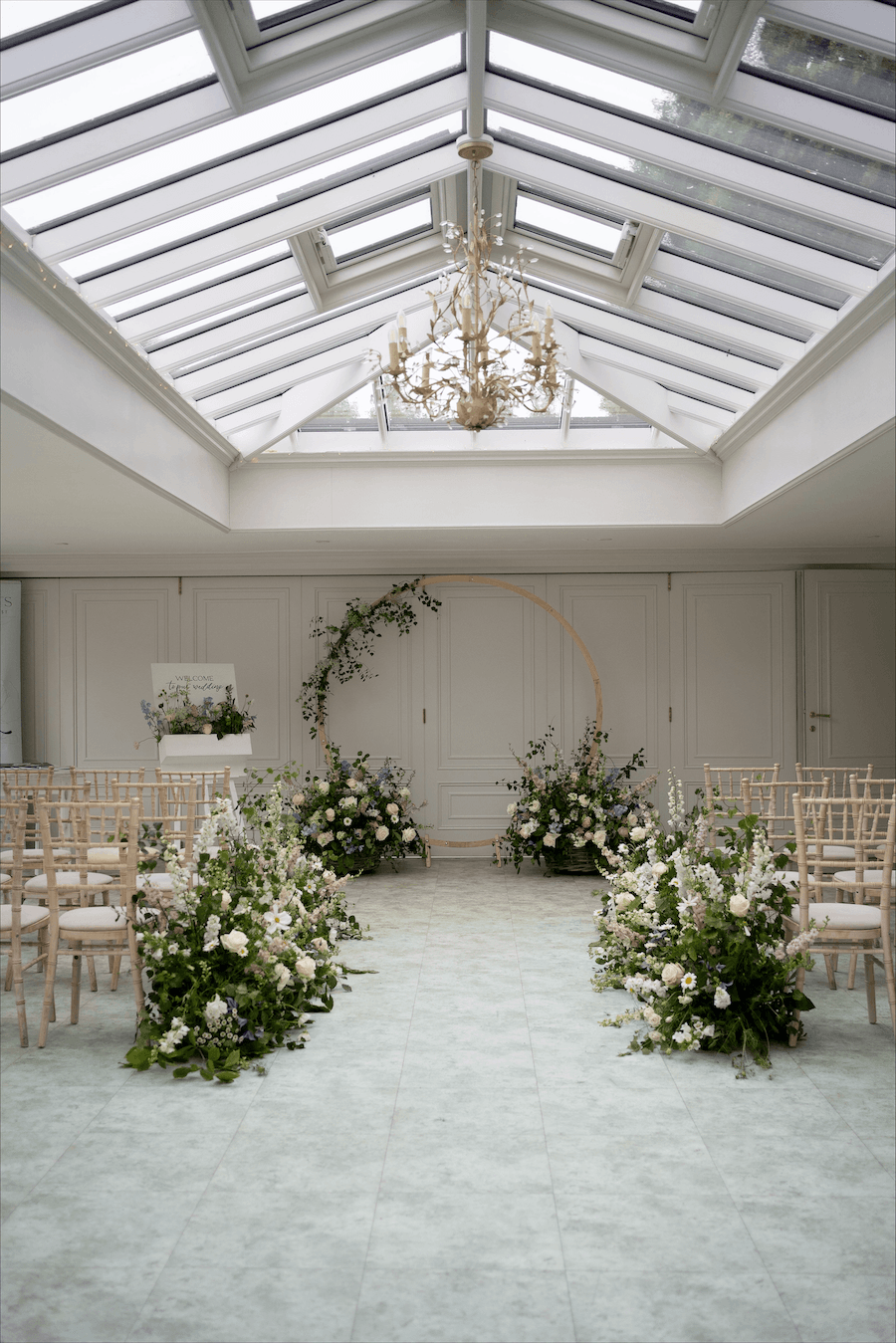 An elegant wedding ceremony setup with rows of beige chairs, white and purple floral arrangements along the aisle, a circular floral arch at the front, and a glass ceiling with a chandelier overhead.