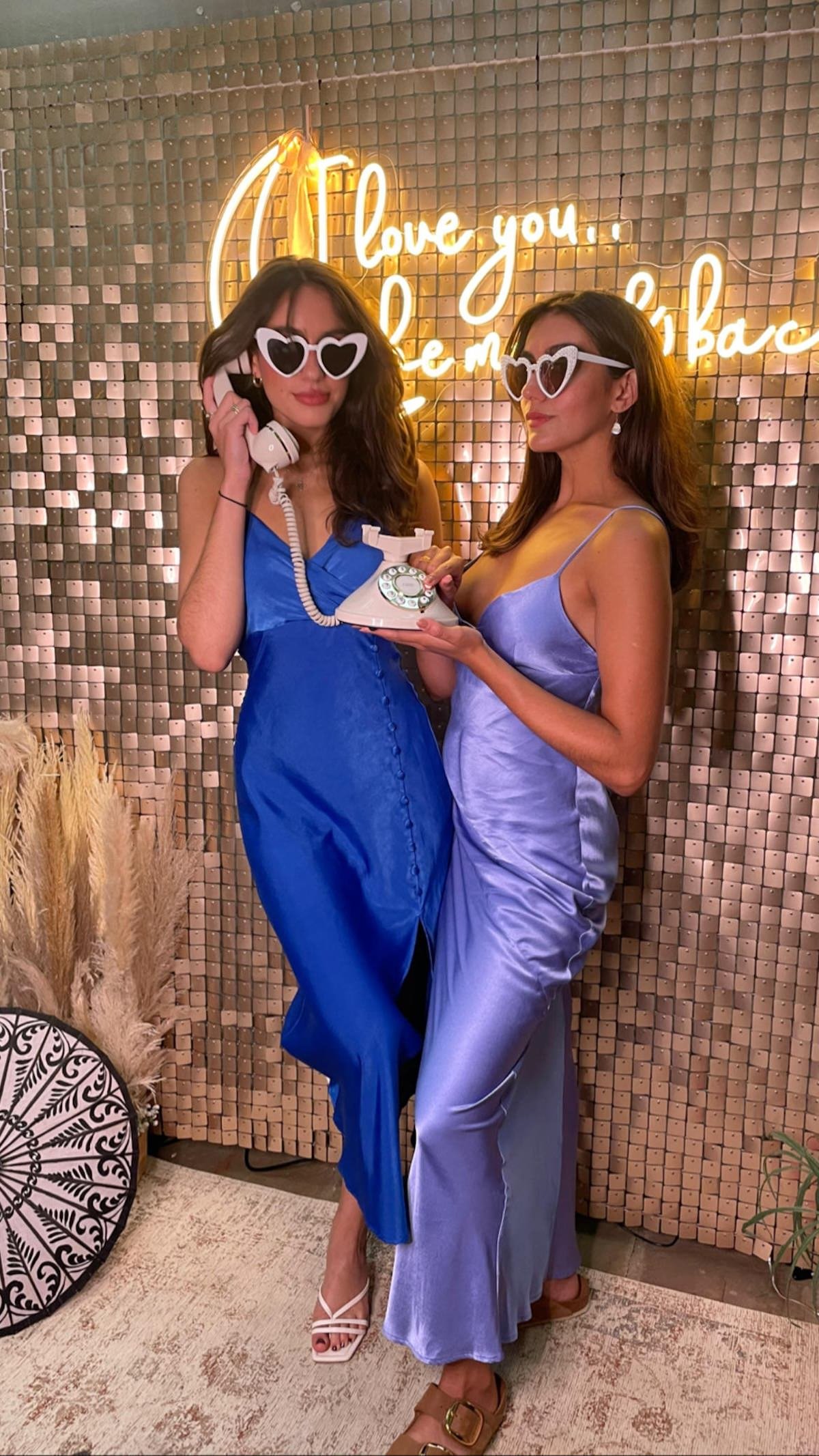 Two women wearing satin dresses and heart-shaped sunglasses stand in front of a wall with a neon sign that says "Love you. Be mine." They are holding a vintage rotary telephone. One woman is wearing a blue dress with white strappy heels, while the ot