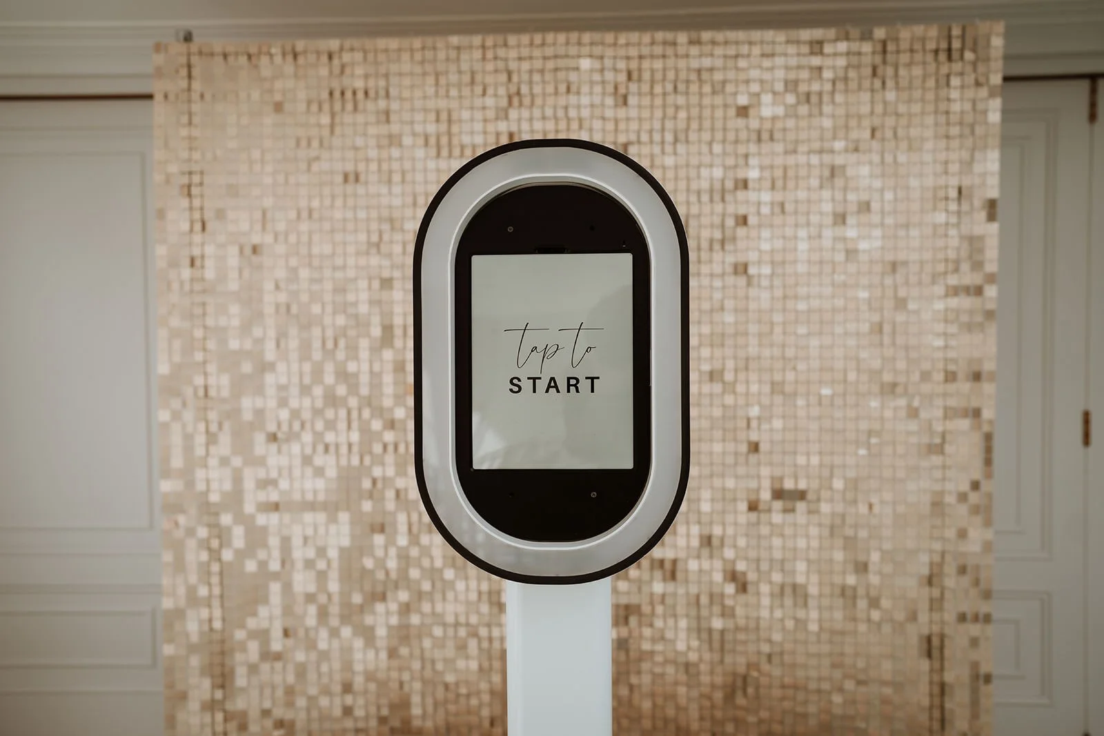 Touch screen photo booth with the text 'Tap to Start' on a beige tiled wall background.