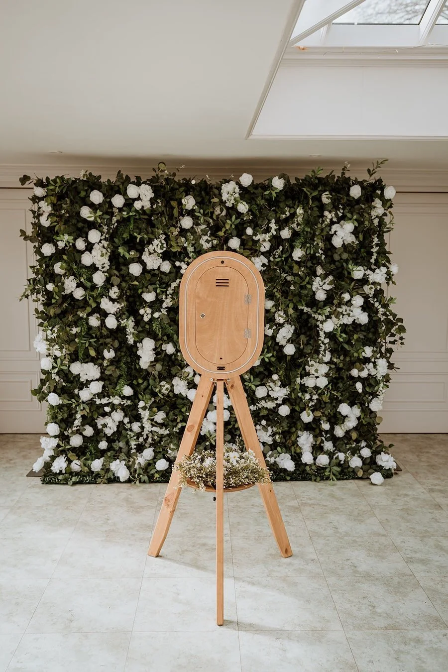 A wooden photo booth with a floral backdrop of white flowers and green leaves, positioned indoors on a tiled floor.