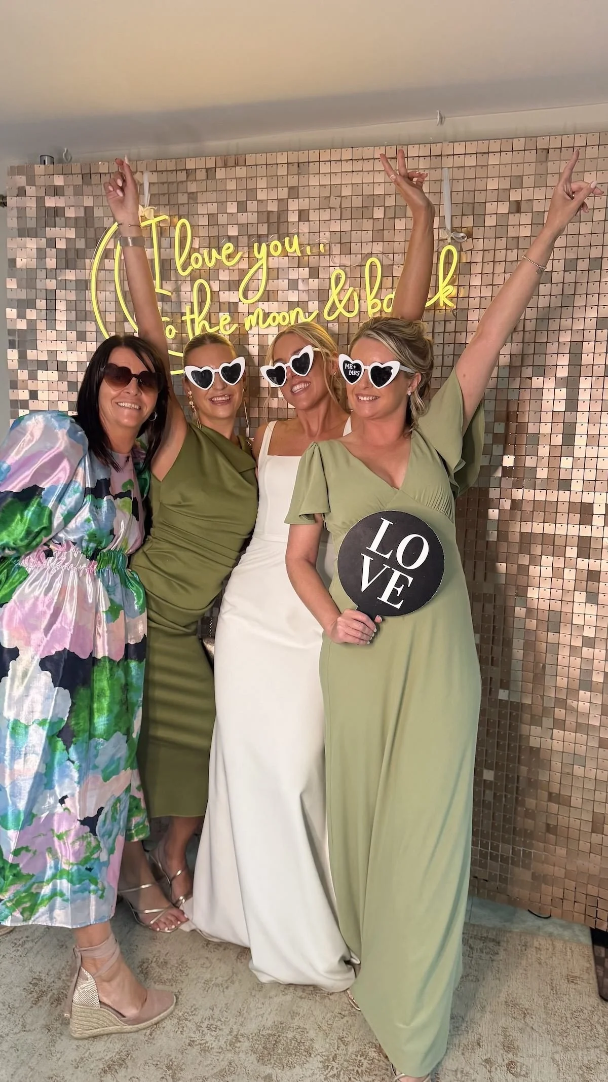 Four women dressed in colorful and elegant dresses, wearing sunglasses, standing together in front of a glittering wall with neon sign that reads 'Love you... to the moon & back.' One woman is holding a sign that says 'LOVE,' and all are celebrating 