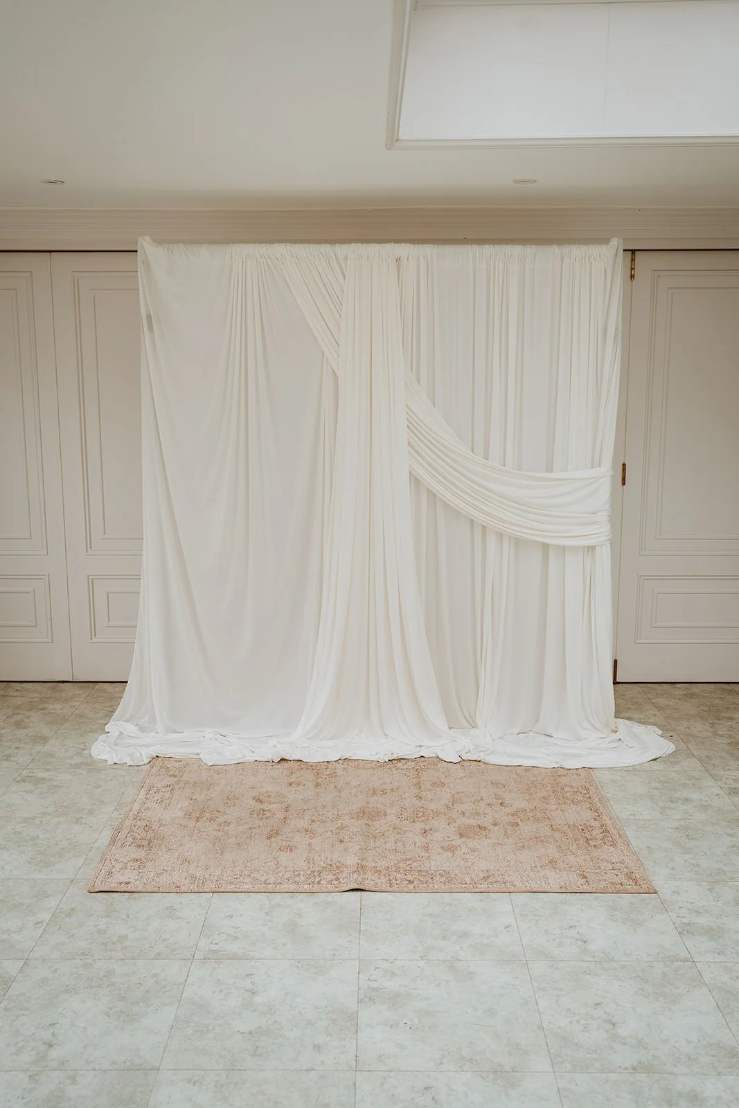 White fabric draped as a backdrop with a gathered section on the right, set against a wall with white paneling, on a beige rug in a room with tiled floor.