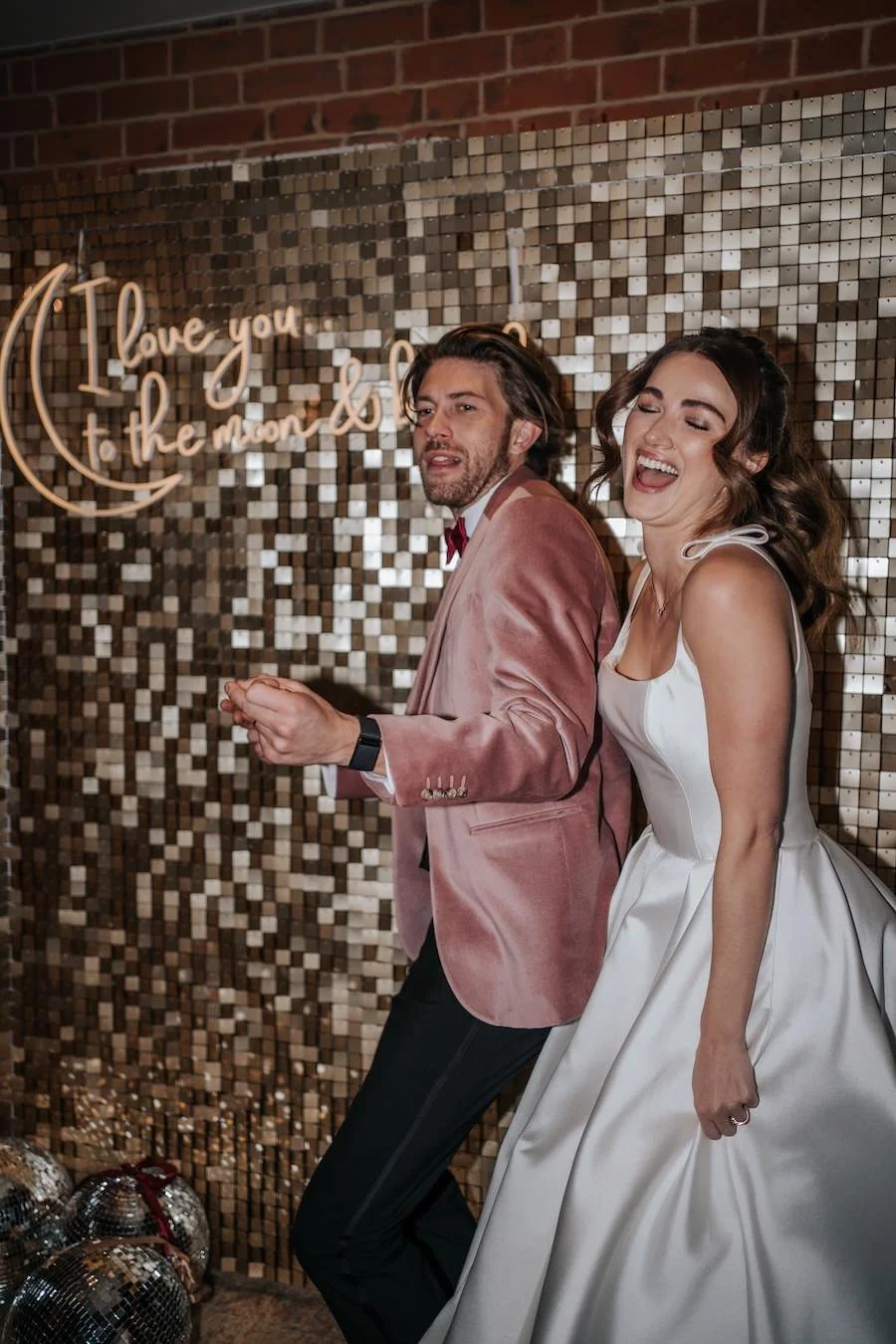 A bride and groom dancing at their wedding reception, with a gold tiled wall and a neon sign saying "I love you to the moon and back" in the background.