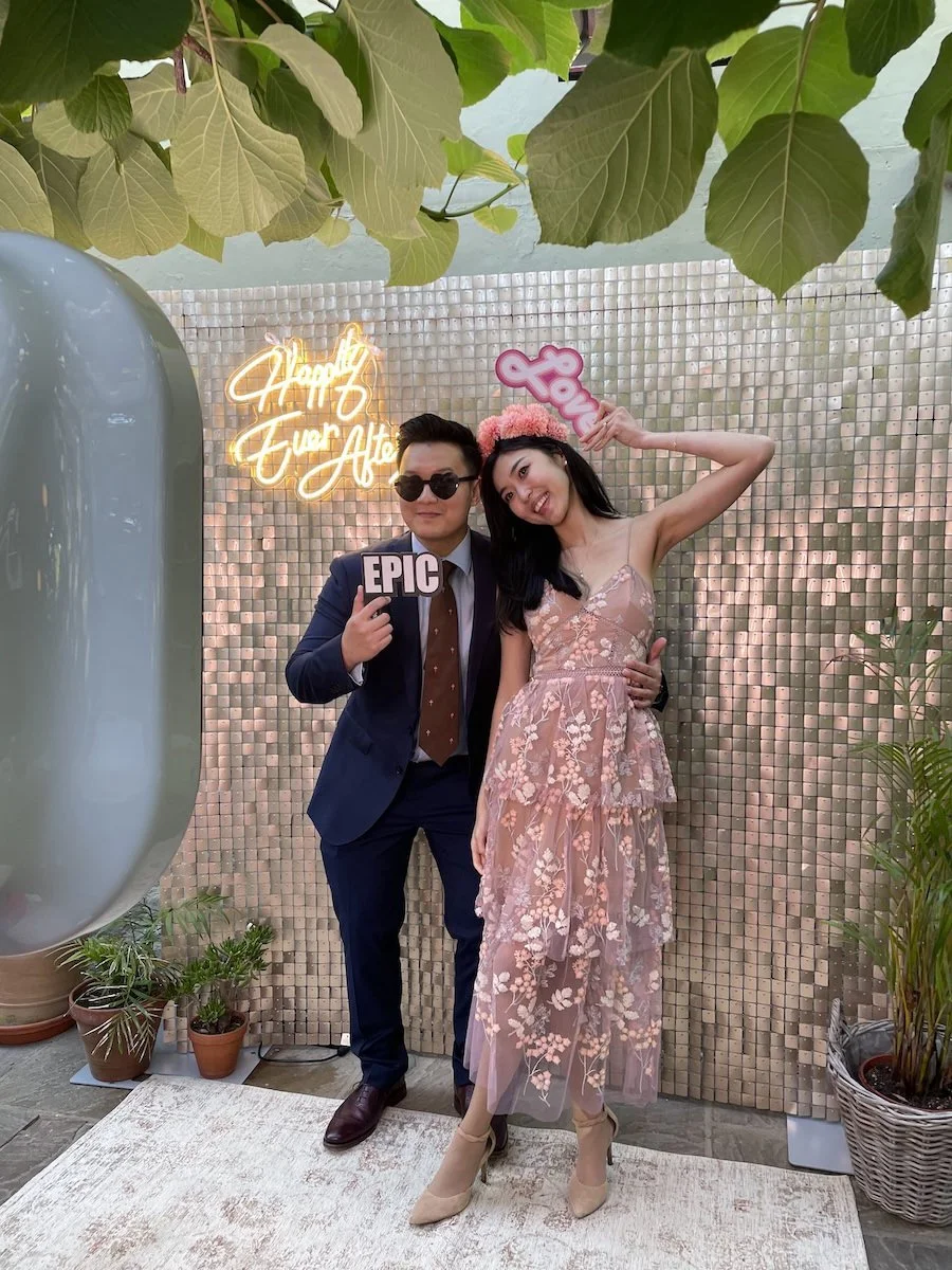 A man in a suit and sunglasses and a woman in a pink floral dress with a pink floral headband pose together in front of a decorated backdrop, holding signs reading 'EPIC' and 'Love,' at a celebration or event.