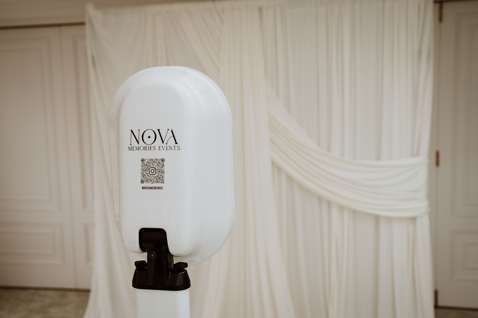 Photo booth with 'NOVA Memories Events' logo, QR code, and Instagram handle in front of draped elegant wedding decorations.