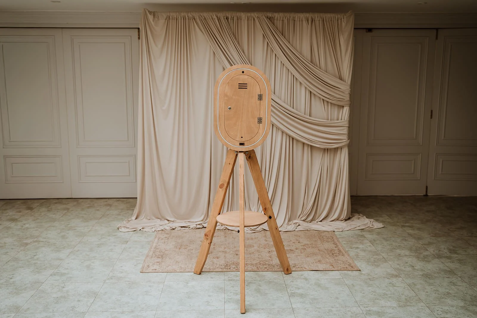 A photo booth mounted on a wooden tripod stand in front of beige curtains with draped detailing, on a tiled floor with a small rug.