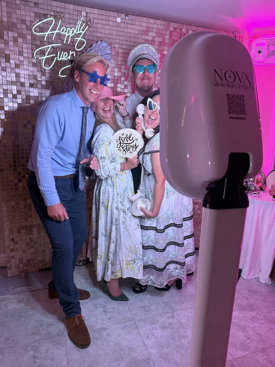 Group of five friends at a celebration party taking a photo with a photo booth. They are wearing funny glasses and holding signs, with a neon sign that says 'Happily Ever' in the background and pink lighting.