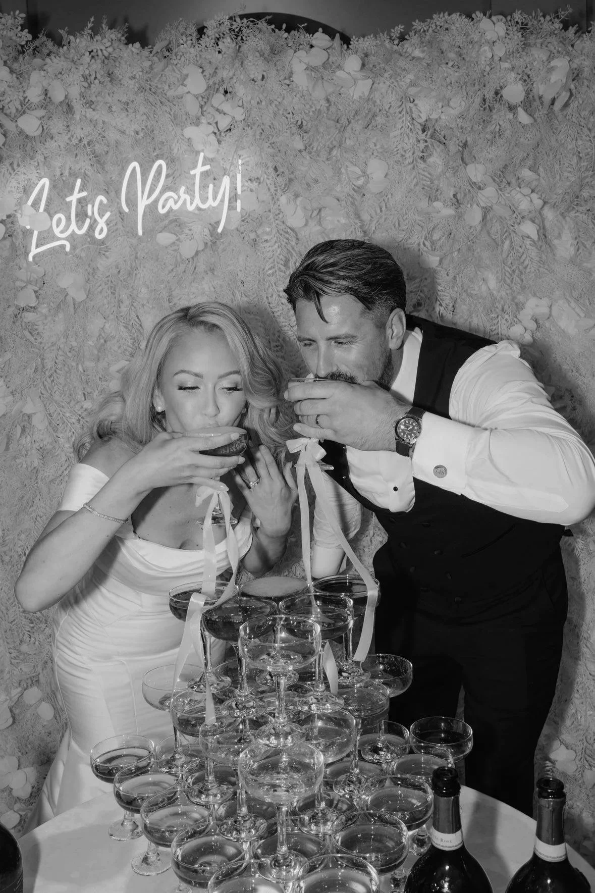 A black and white photo of a man and woman at a celebration, pouring drinks into glasses stacked on a table, with flowers and the phrase 'Let's Party!' in the background.