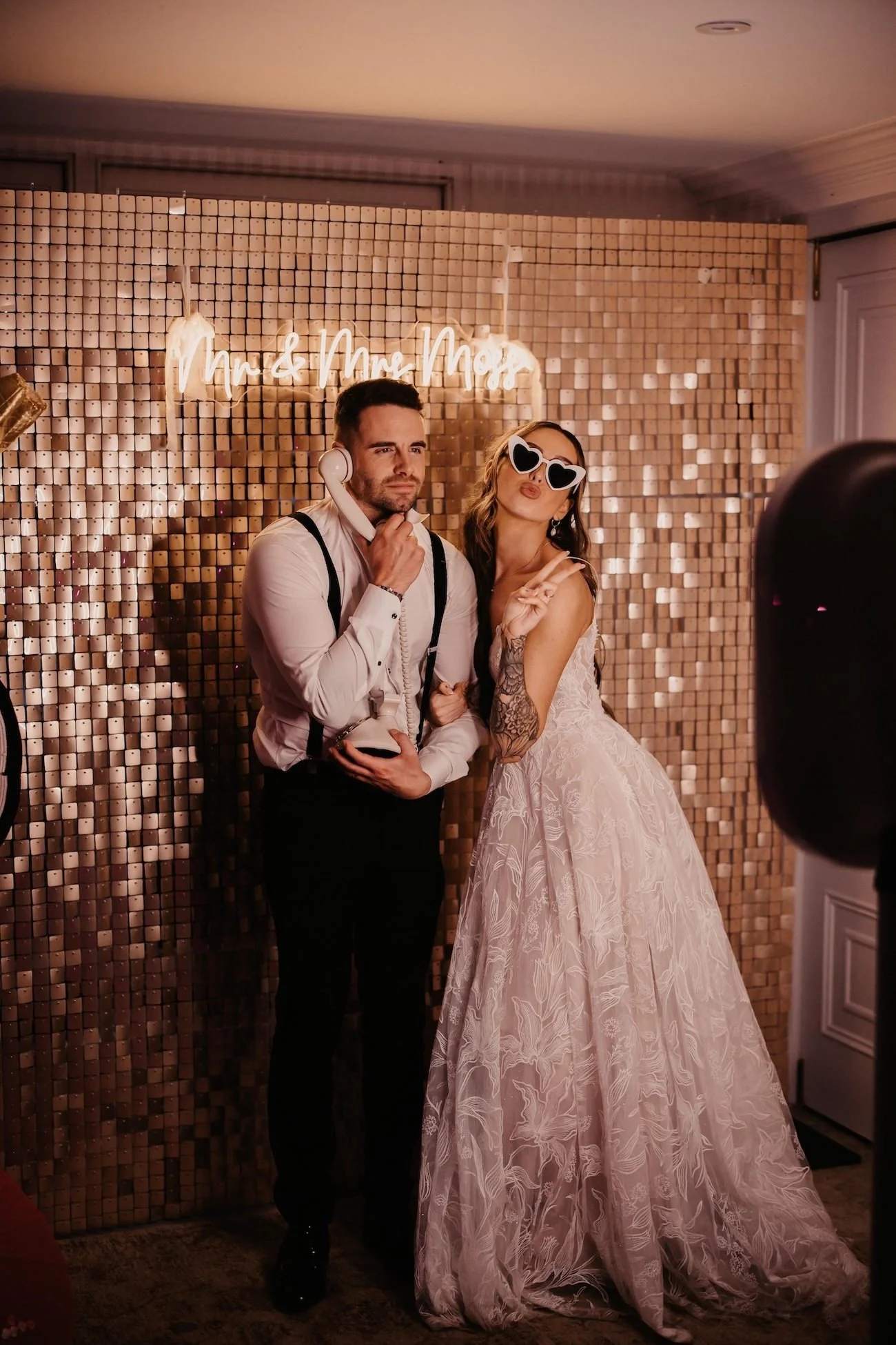 A man with headphones and suspenders and a woman in a wedding dress with sunglasses making peace sign pose at a party with a copper-colored backdrop and a neon sign that reads 'Mr & Mrs Moe'.