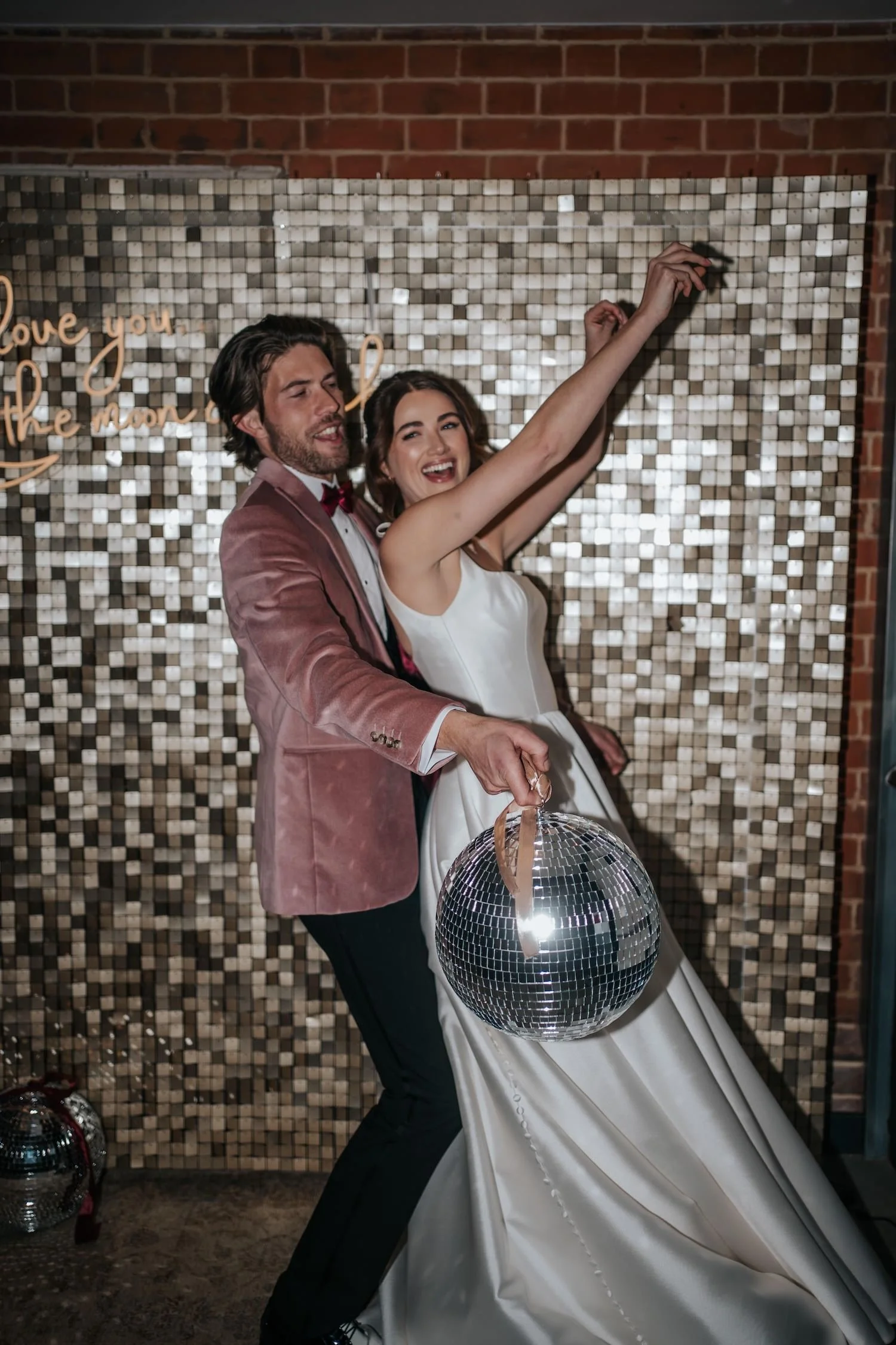 A man and woman dancing at a celebration, with the woman holding a disco ball, in front of a sparkling backdrop with a brick wall above.