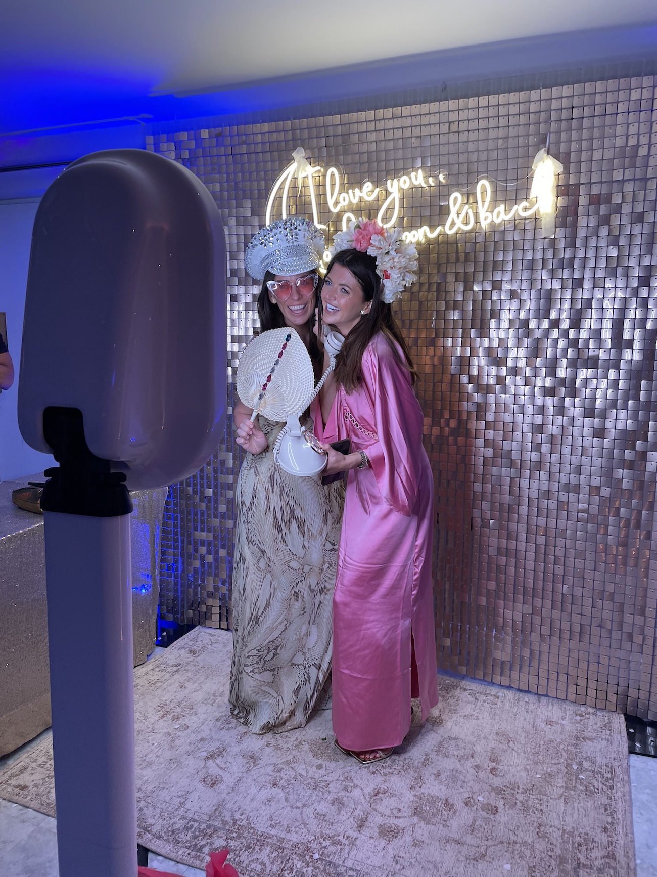 Two women wearing colorful, festive outfits and floral headpieces, smiling and posing together at a party with a metallic backdrop and neon sign that reads 'Love you, who we are'.