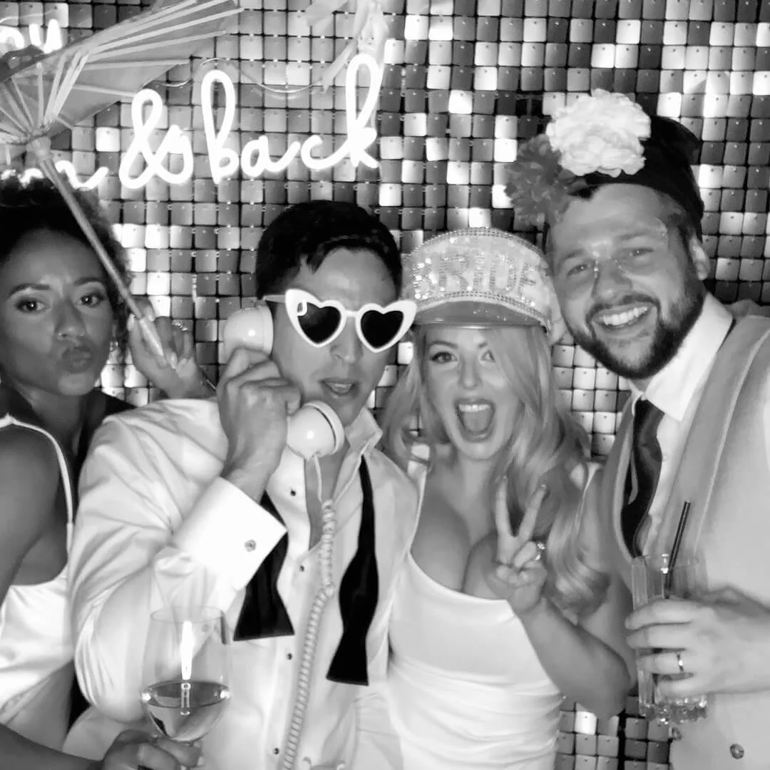 Group of four people celebrating at a party, with a sign that says 'I'll be back' in the background. They are wearing fun accessories, including sunglasses, a hat, and a flower headpiece, and they are holding drinks.