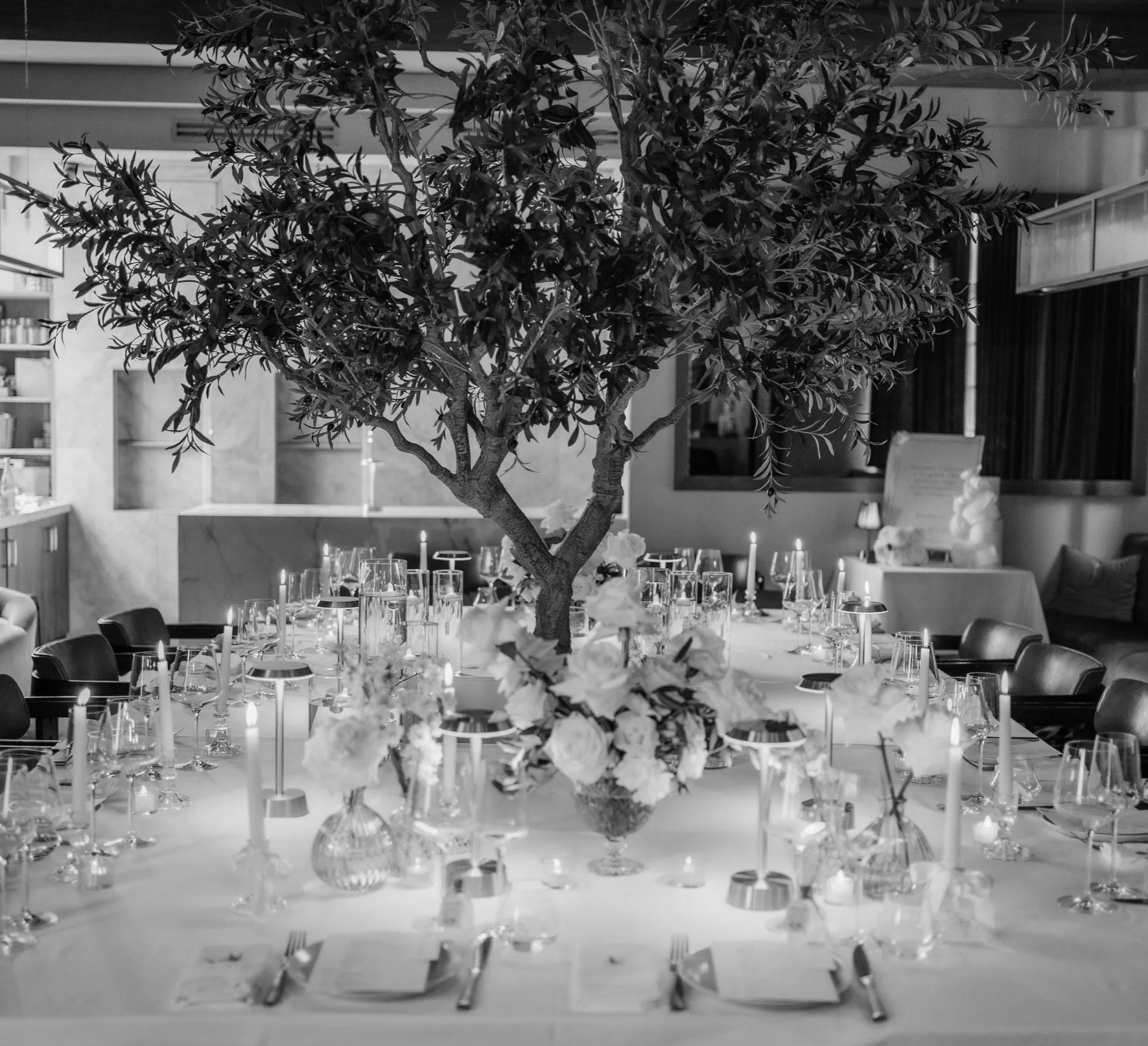 A large indoor space decorated for a formal event with a tree in the center of the dining table, surrounded by glassware and floral arrangements.