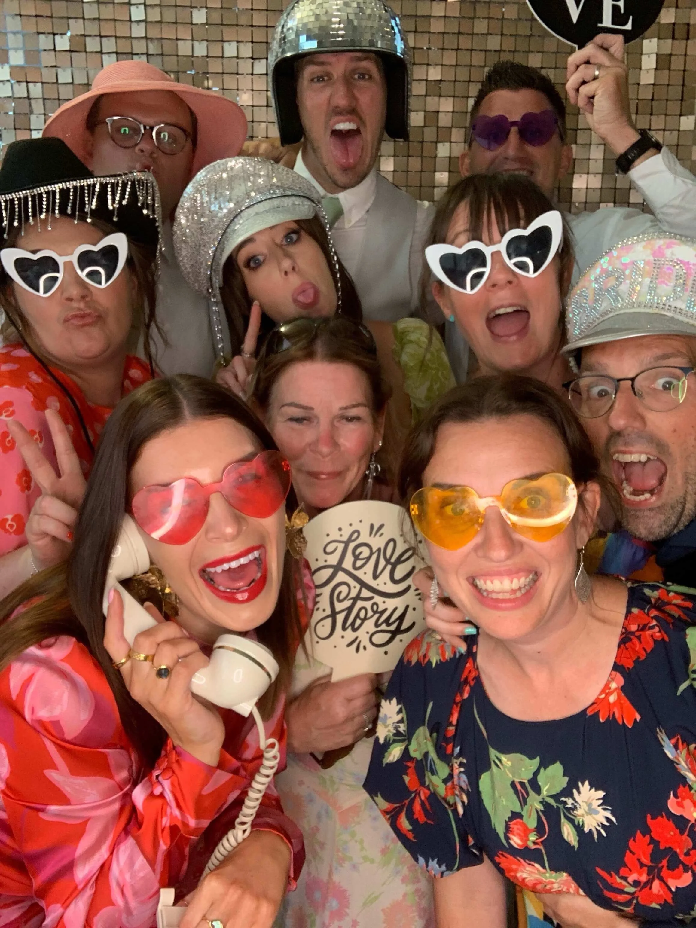 A group of people in a party, wearing colorful and shiny accessories, sunglasses, hats, and holding a sign that says 'Love Story' with a mosaic background.