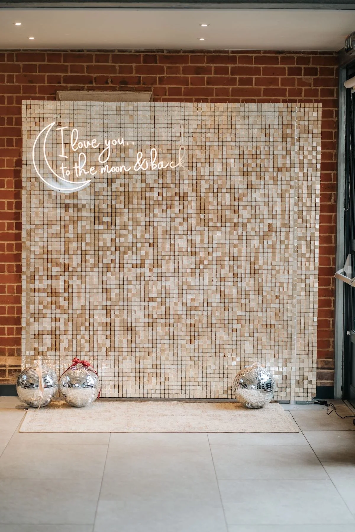 Decorative backdrop with a neon sign that says "I love you to the moon & back" and three silver disco balls at the base, set against a brick wall.