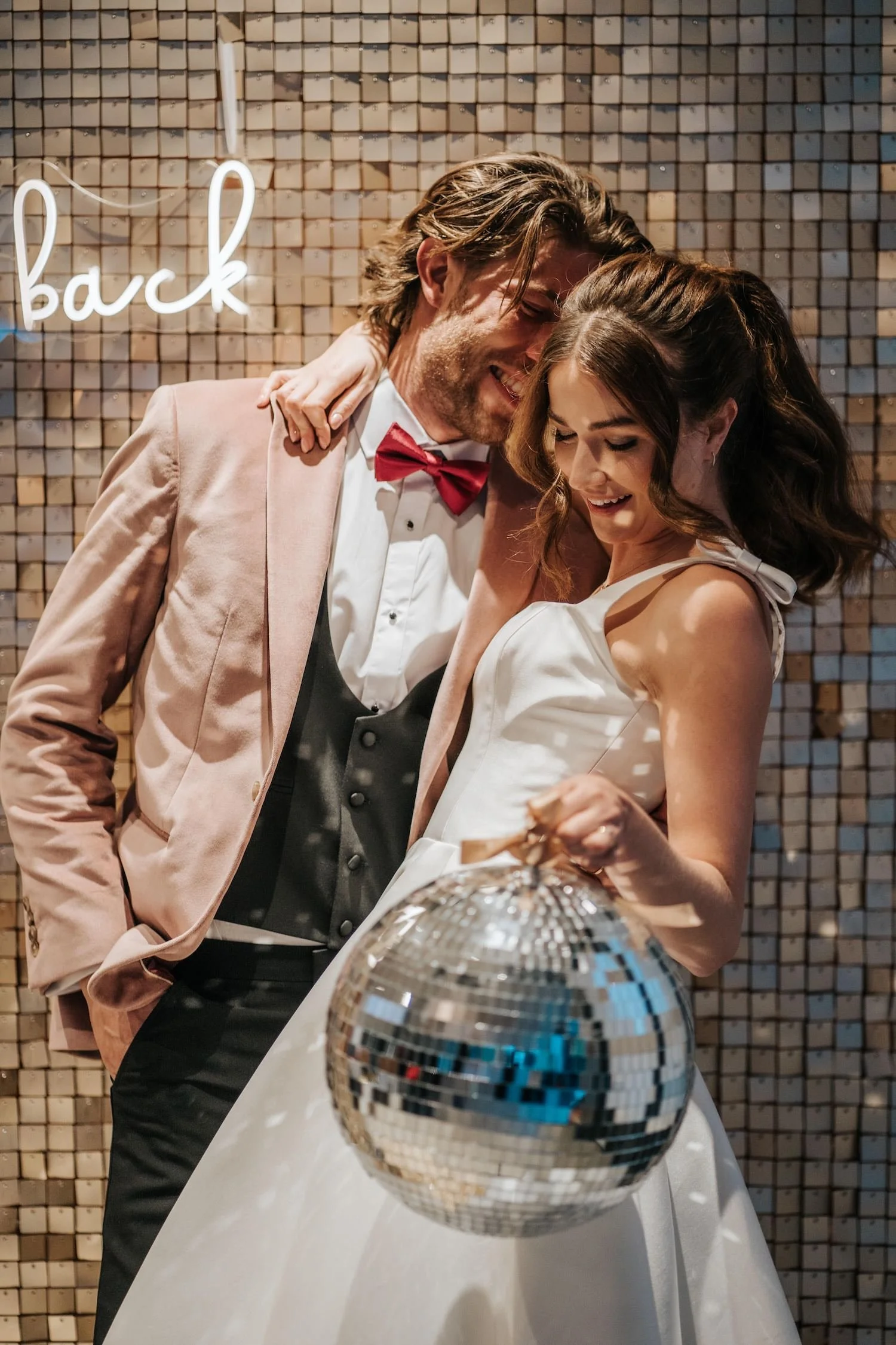 A man and woman dressed in formal attire smiling and dancing close together at a party, with a disco ball and a mosaic tile wall in the background, and a neon sign that says "back."