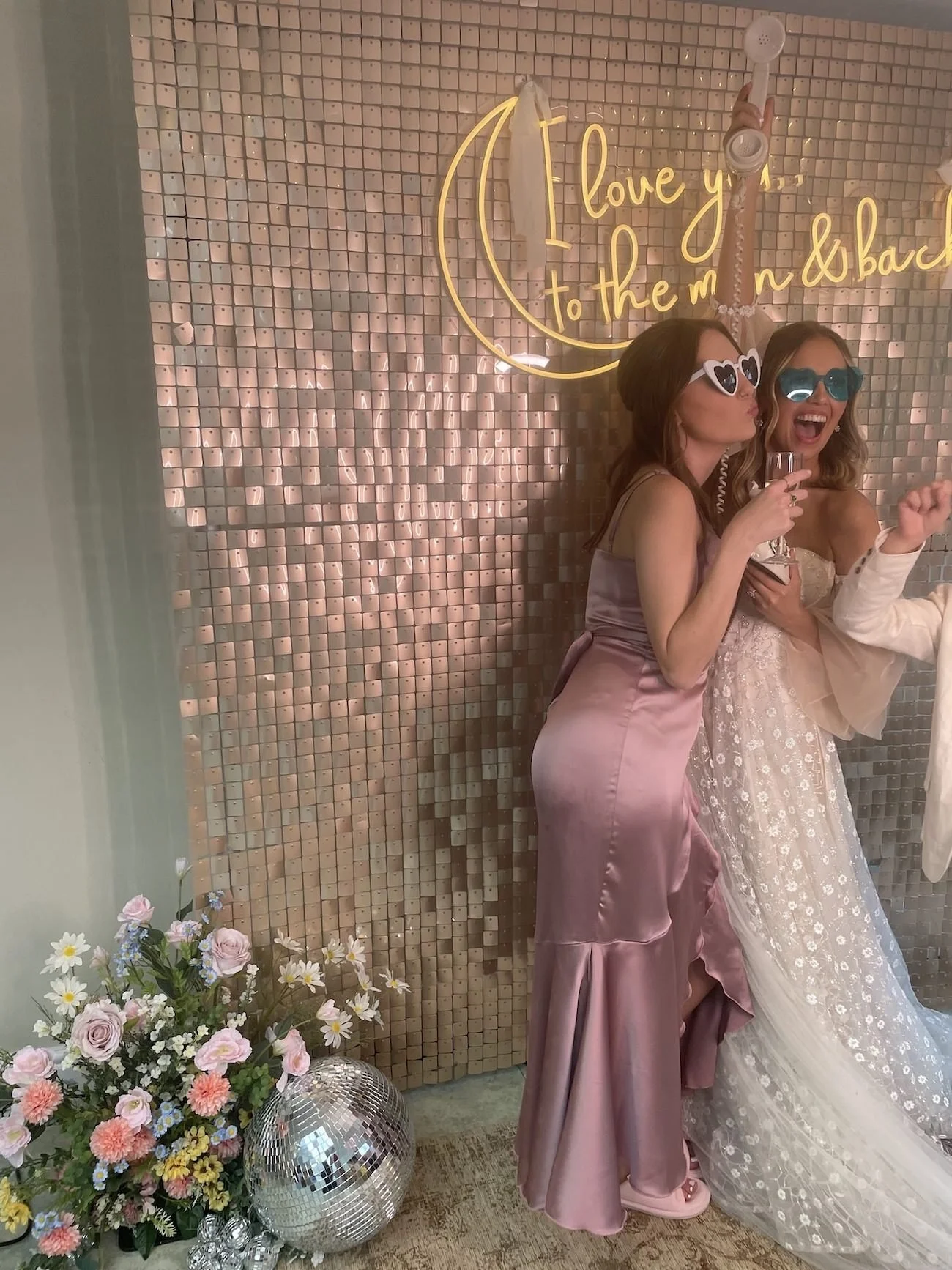 Two women in formal dresses wearing sunglasses, standing in front of a pink mosaic wall with illuminated yellow text. One woman is wearing a satin pink gown, and the other a white wedding dress. There is a floral bouquet and a disco ball on the floor