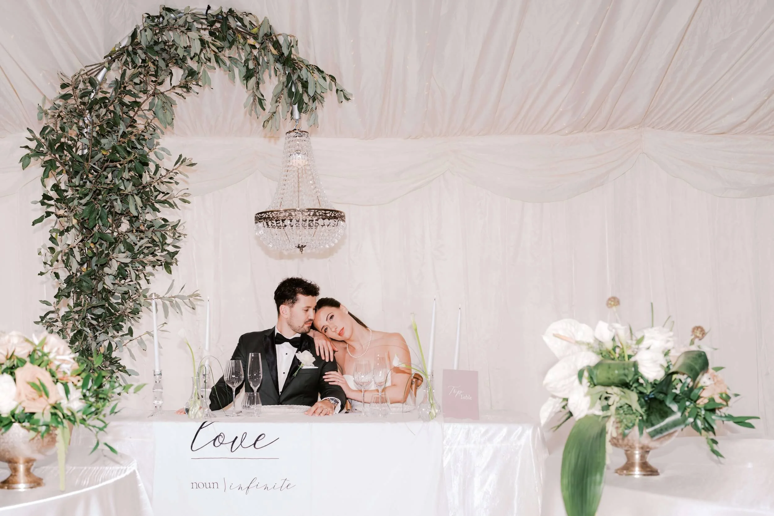 A bride and groom sitting at a decorated wedding reception table with floral centerpieces, greenery, and a chandelier above. The table has a sign that says 'love' and elegant glassware.