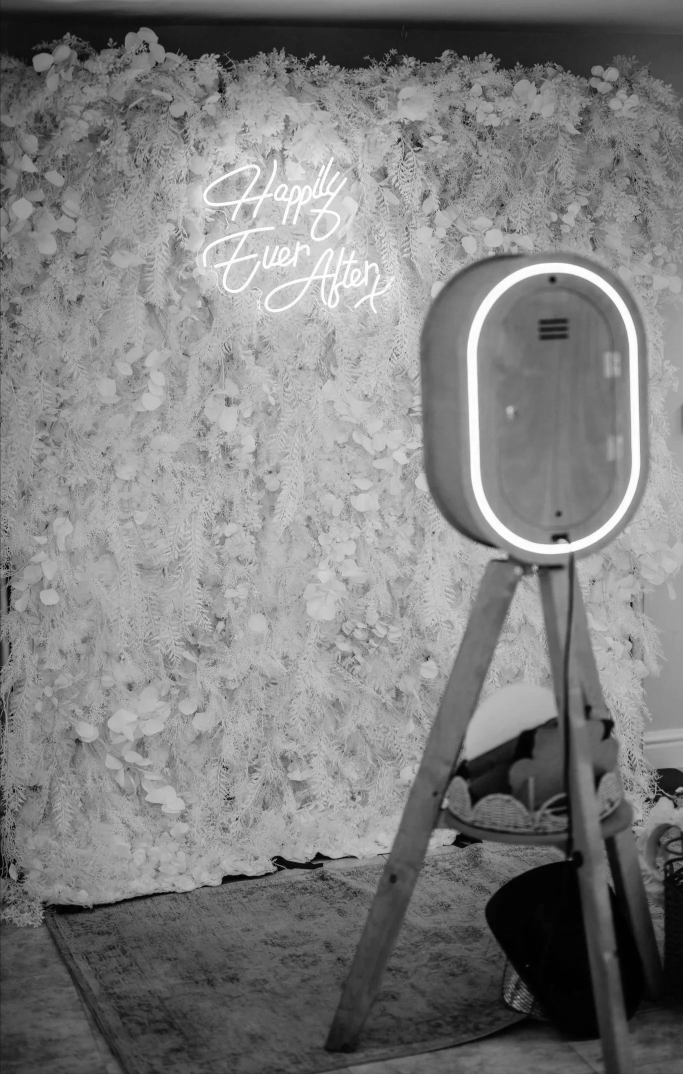 Black and white photograph of a floral backdrop with a neon sign that reads 'Happily Ever After,' a camera on a tripod, a chair, and a basket on the floor.