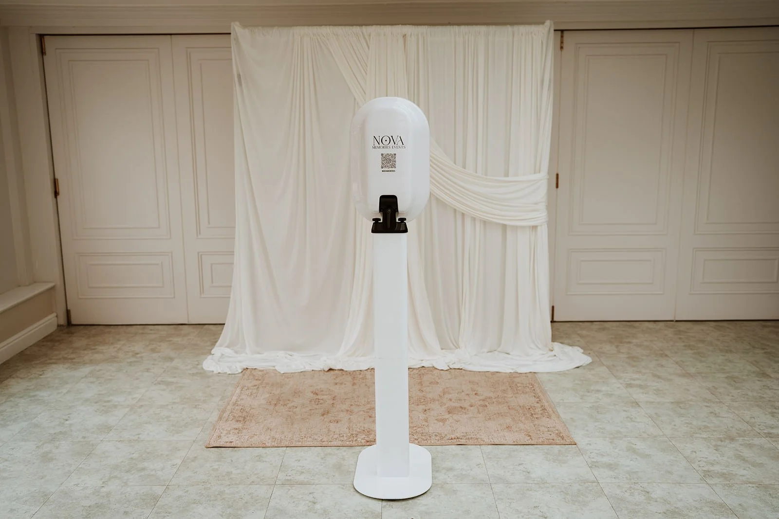Photo booth with a white stand and a large bottle labeled 'NOVA Memories & Events,' in front of cream-colored draped curtains and closed closet doors.